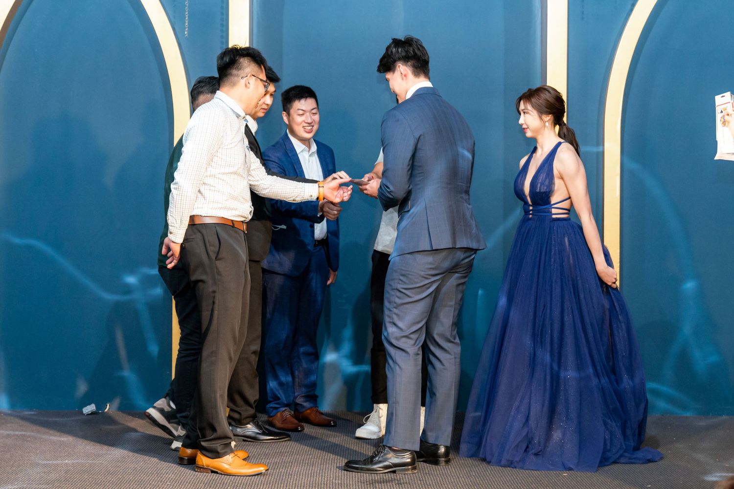 People in formal attire interact at an event against a blue backdrop, with someone wearing an elegant navy blue gown.