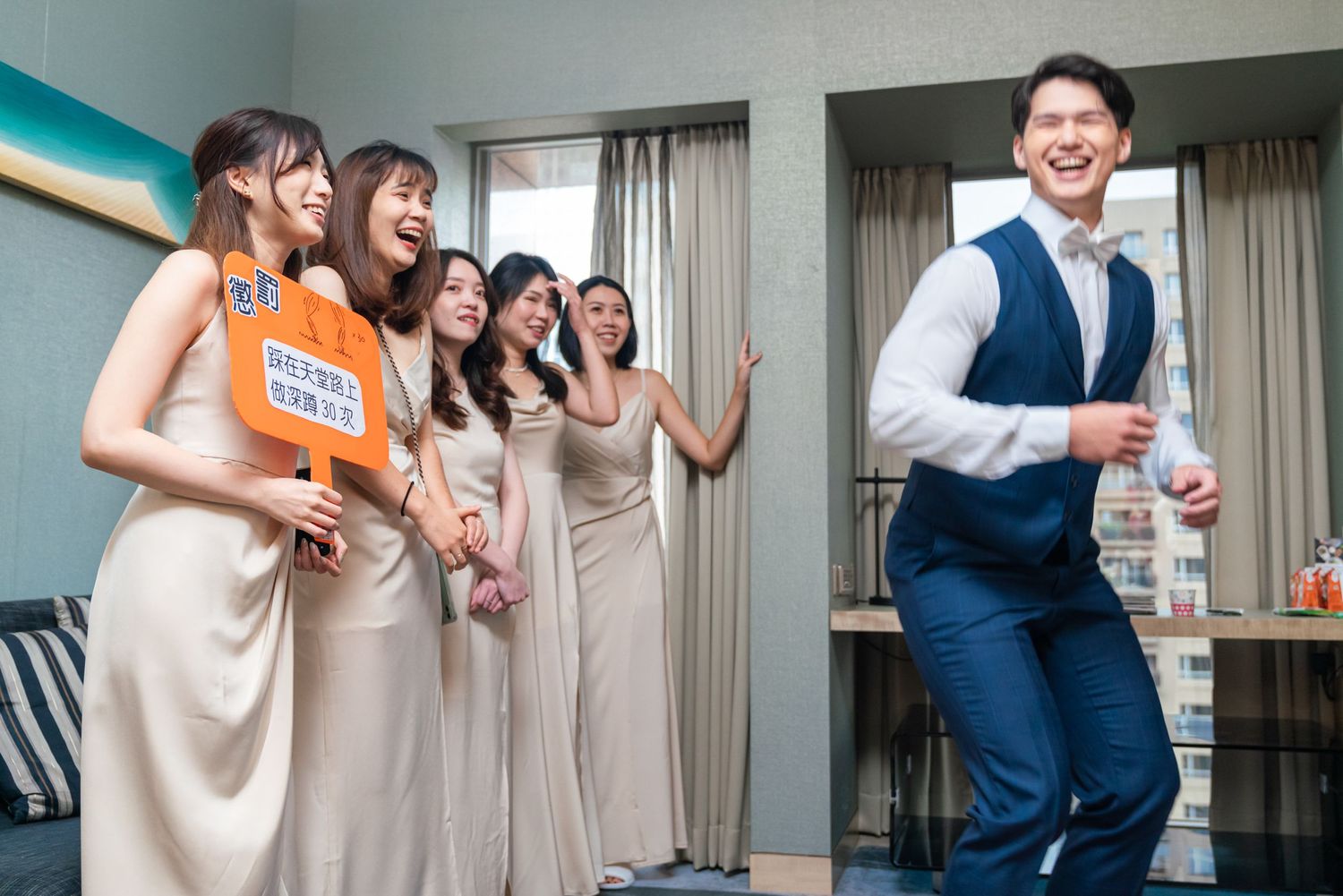 Bridesmaids in champagne dresses laugh together as someone in a blue vest dances nearby at a wedding celebration.