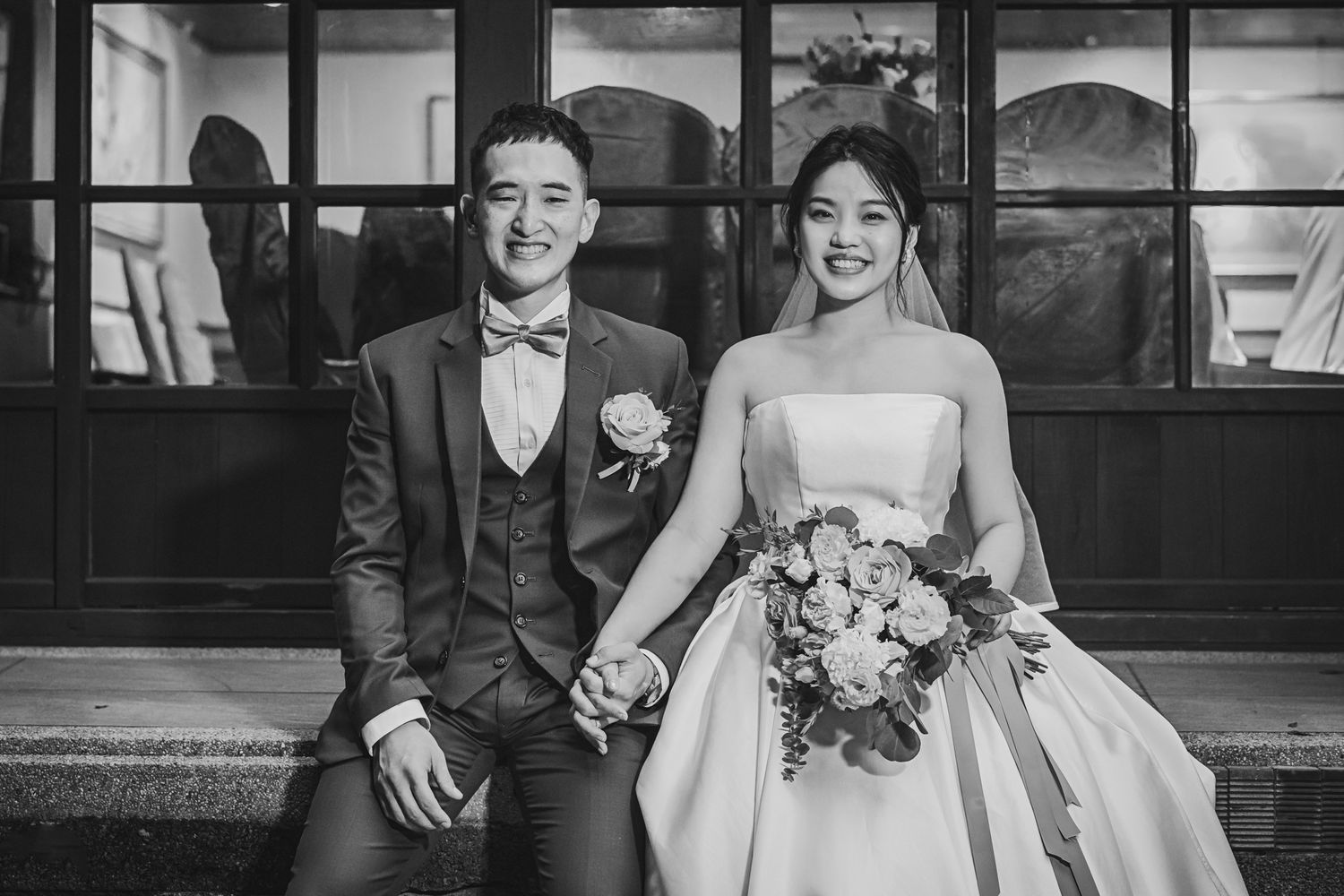 A bride and groom sit together on steps holding hands in an elegant black and white wedding portrait.
