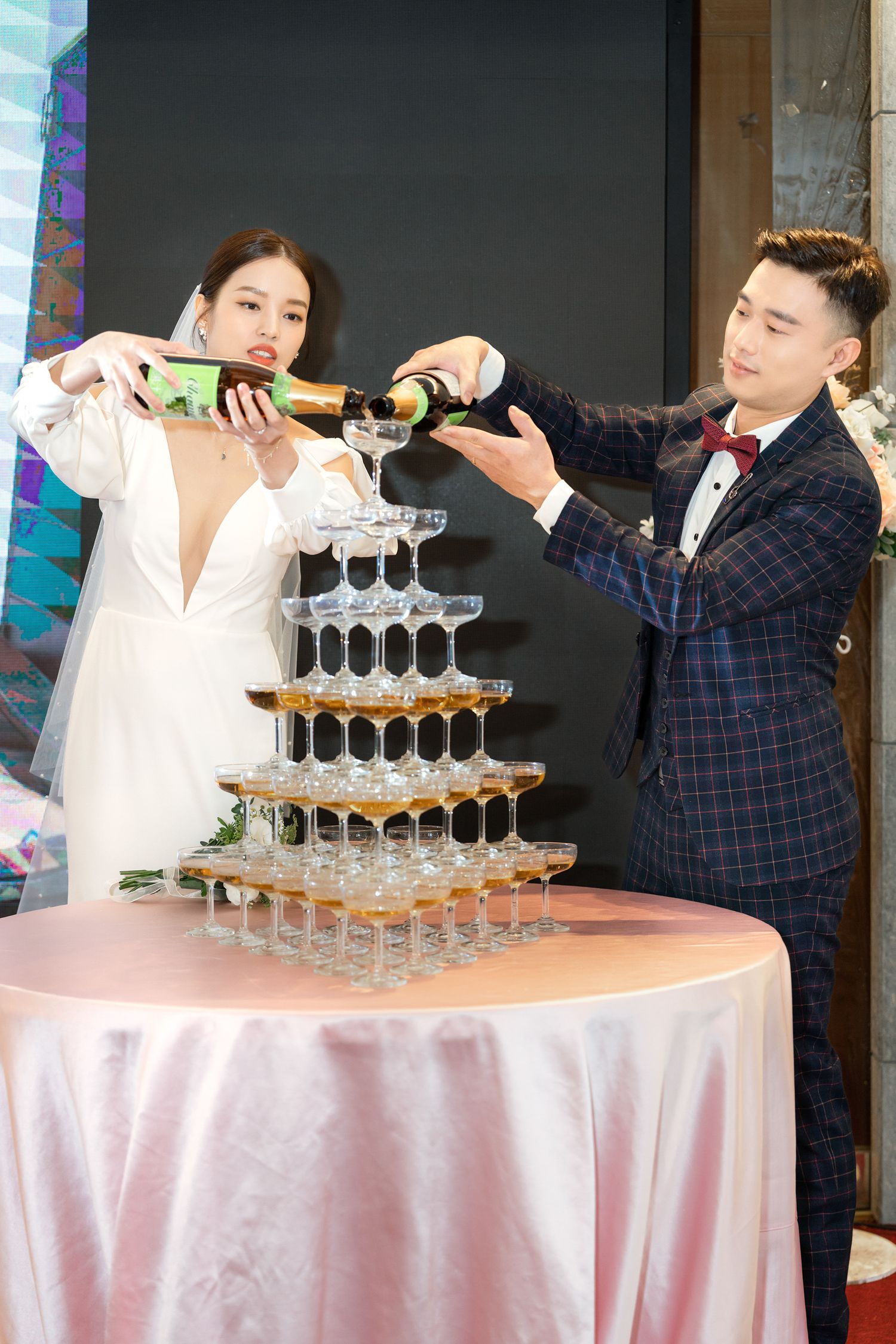 Two people pour champagne into a multi-tiered tower of glasses at an elegant celebration.