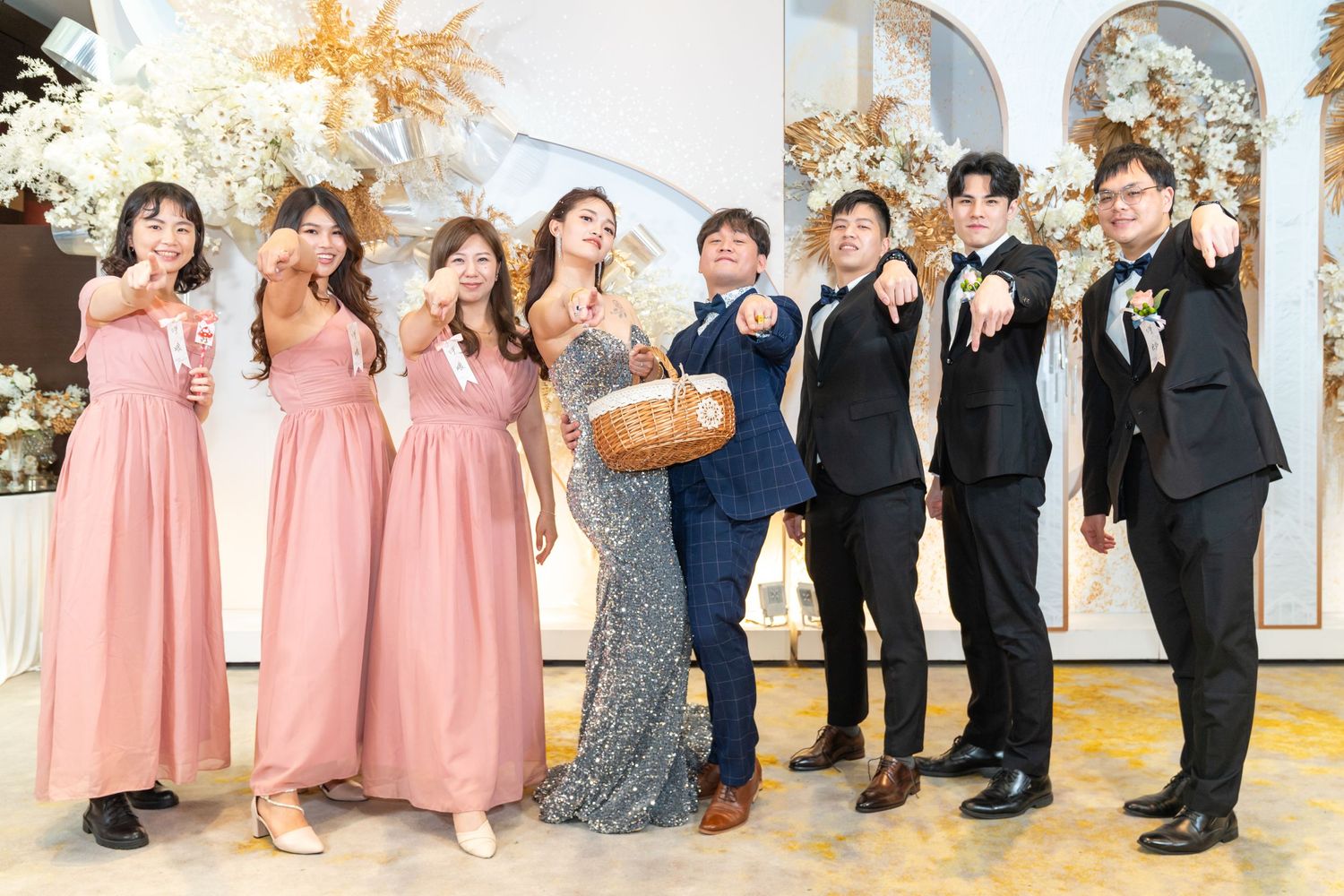 Wedding party in formal attire with bridesmaids in pink dresses and groomsmen in black suits pose together at decorated venue.