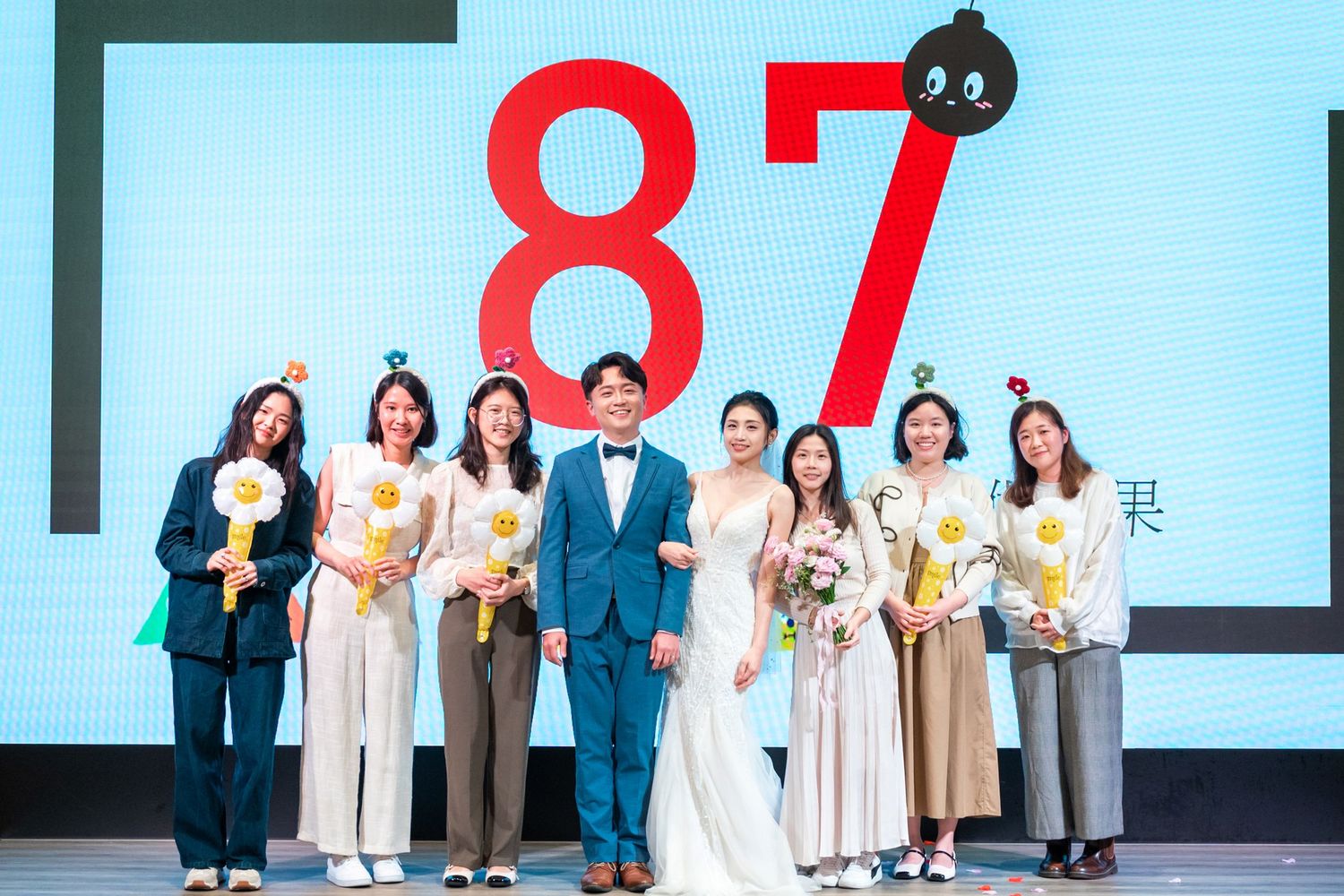A group stands in front of a large blue wall with the number 87 displayed, holding flowers and dressed in formal attire.