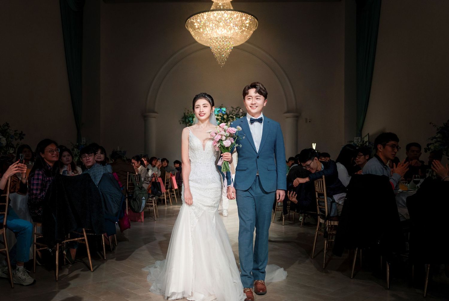 A bride and groom walk down the aisle together in an elegant venue with a crystal chandelier and seated guests.