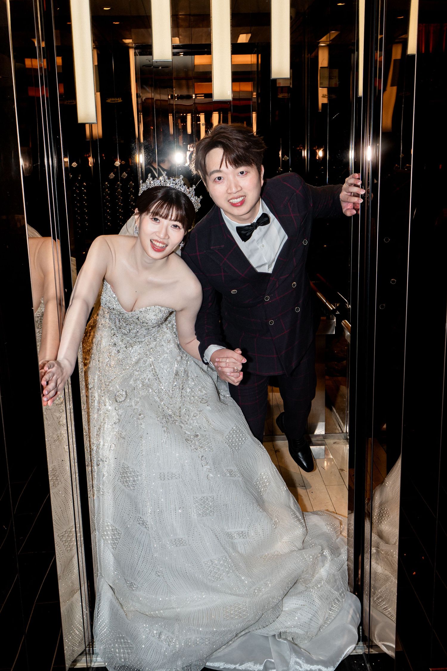 A bride in a silver gown and tiara poses with a groom in a black tuxedo in an elevator at their wedding.