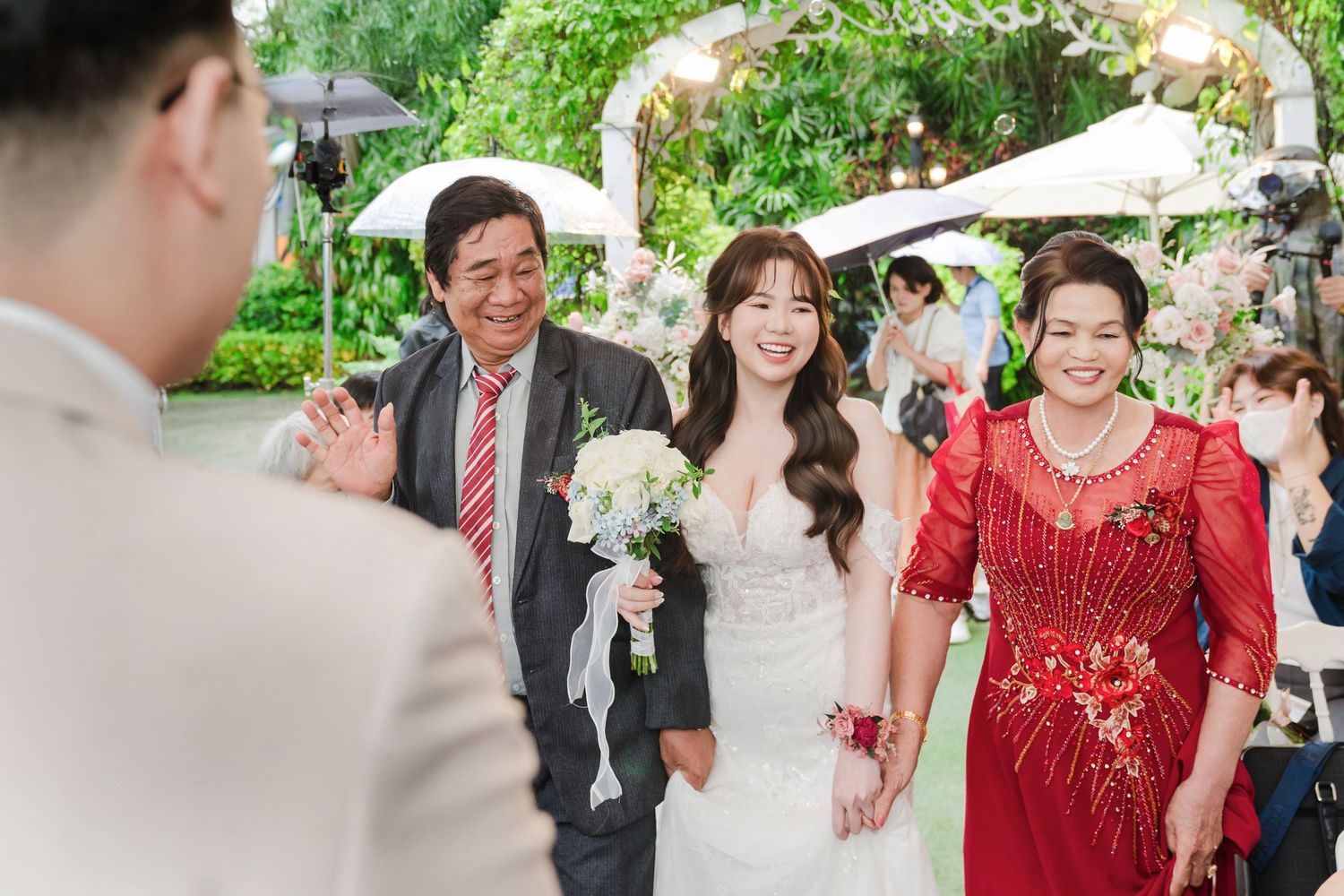 A wedding ceremony procession with the bride in white holding a bouquet walking alongside a woman in an elegant red dress.