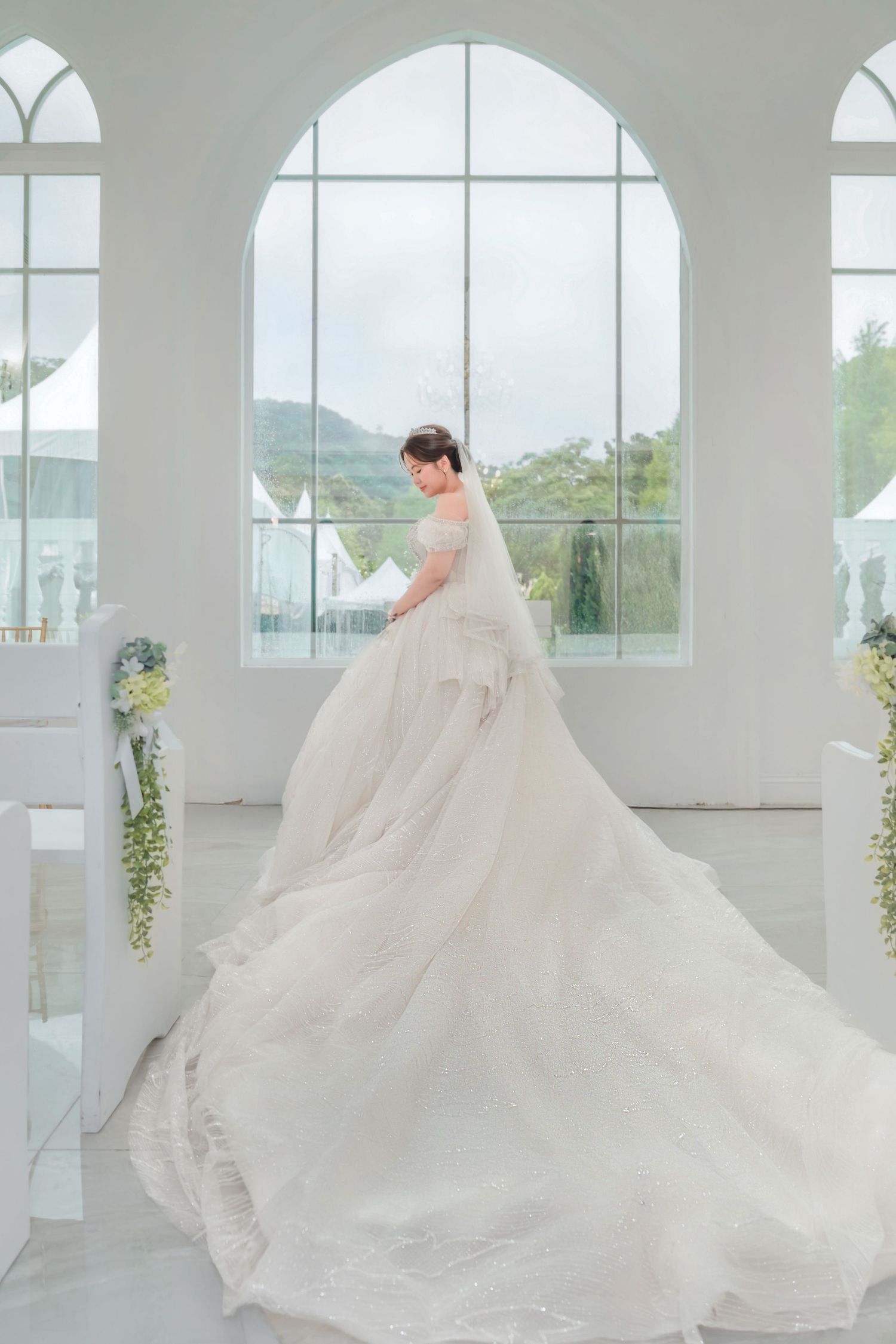 A person in an elegant white wedding gown with flowing train stands in a bright sunlit chapel with arched windows.