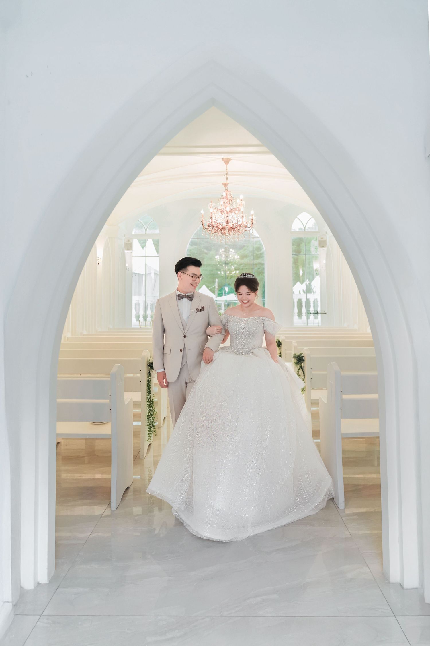 A couple in wedding attire standing under an arched chapel doorway with a crystal chandelier above.