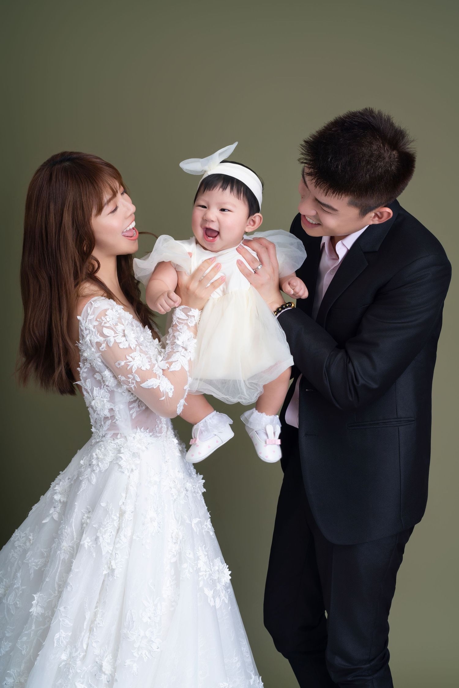 A happy family moment with parents in wedding attire holding their joyful baby in a white dress against a green backdrop.