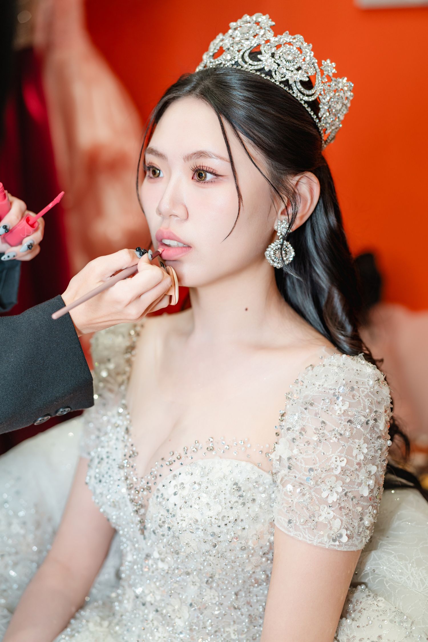A bride wearing an ornate crystal crown and sparkly dress has her makeup applied before her wedding ceremony.