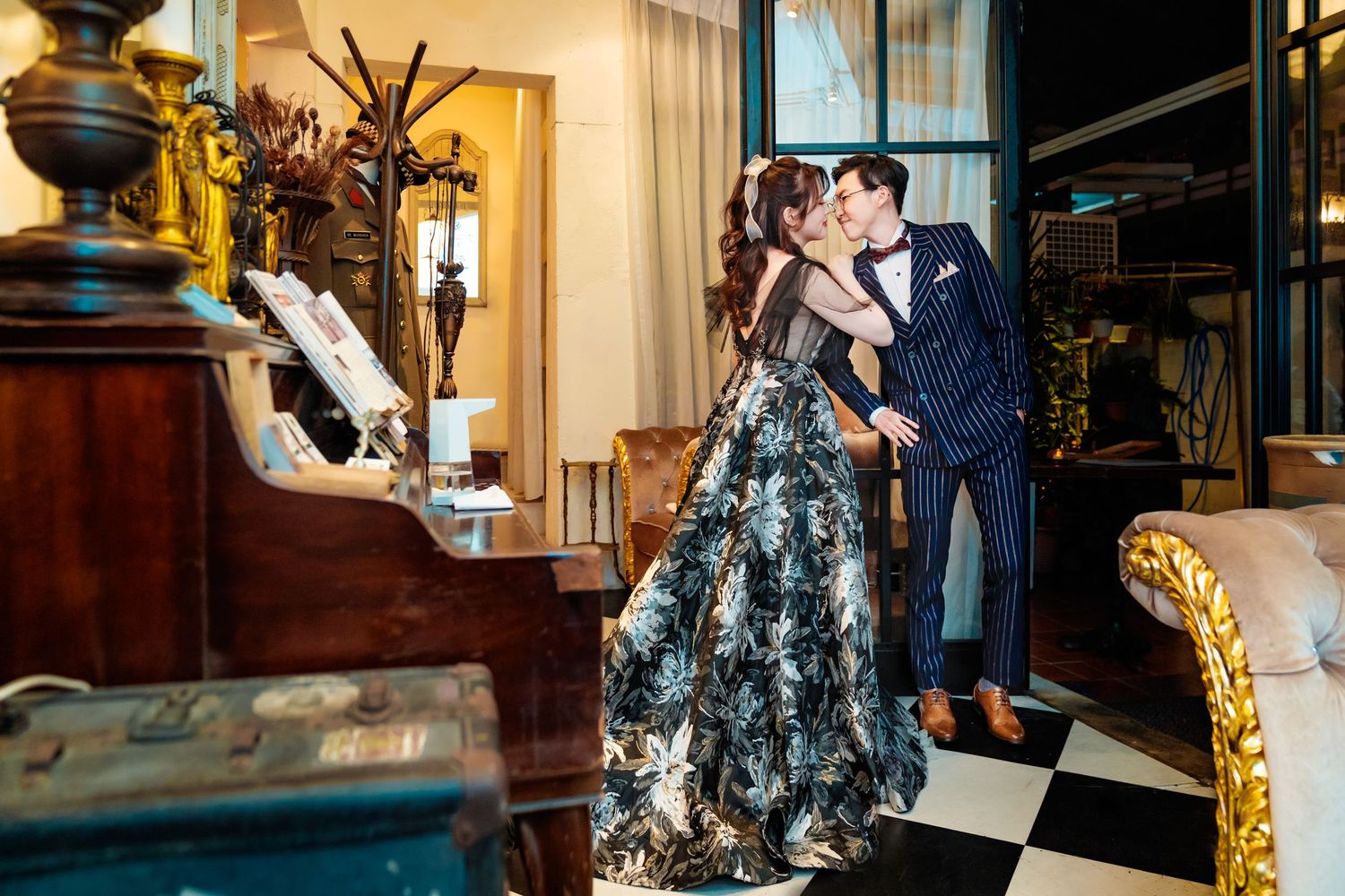 Two people dance intimately in formal attire by an antique piano in an elegant room with checkered floors and warm lighting.