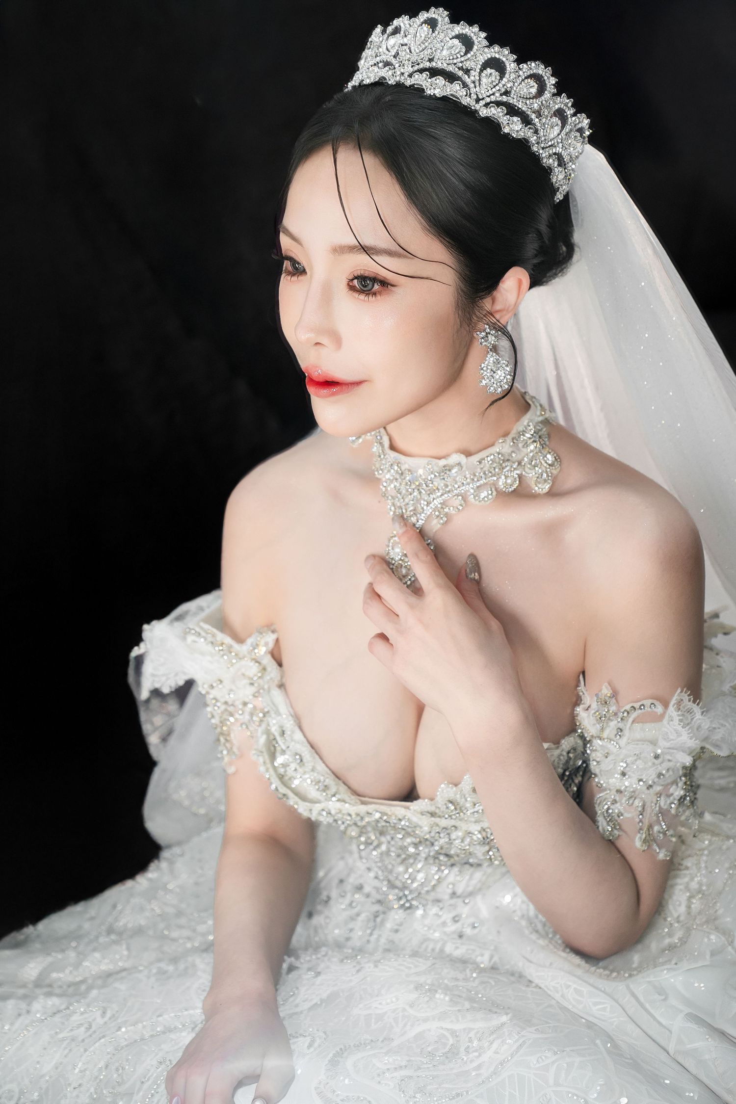 A bride in an ornate white wedding dress with crystal embellishments and veil poses elegantly against a dark background.