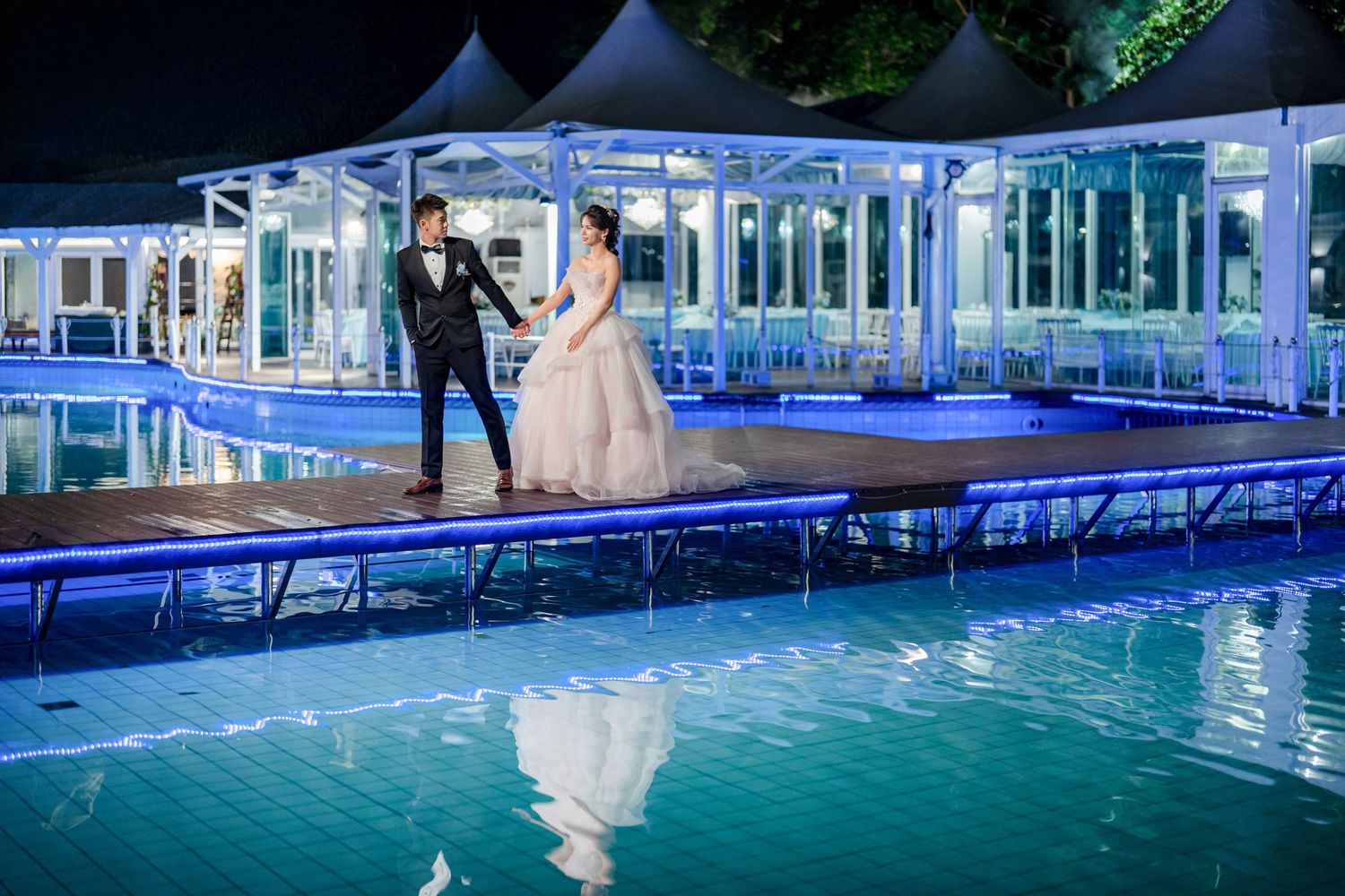 A couple stands on an illuminated walkway by a glowing blue pool with white tents and reflections in the evening.