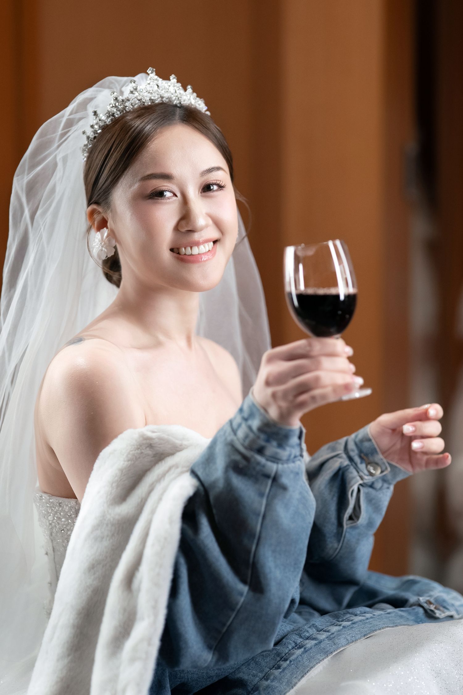 A bride in a white wedding dress and veil holds a glass of red wine while wearing a casual denim jacket.