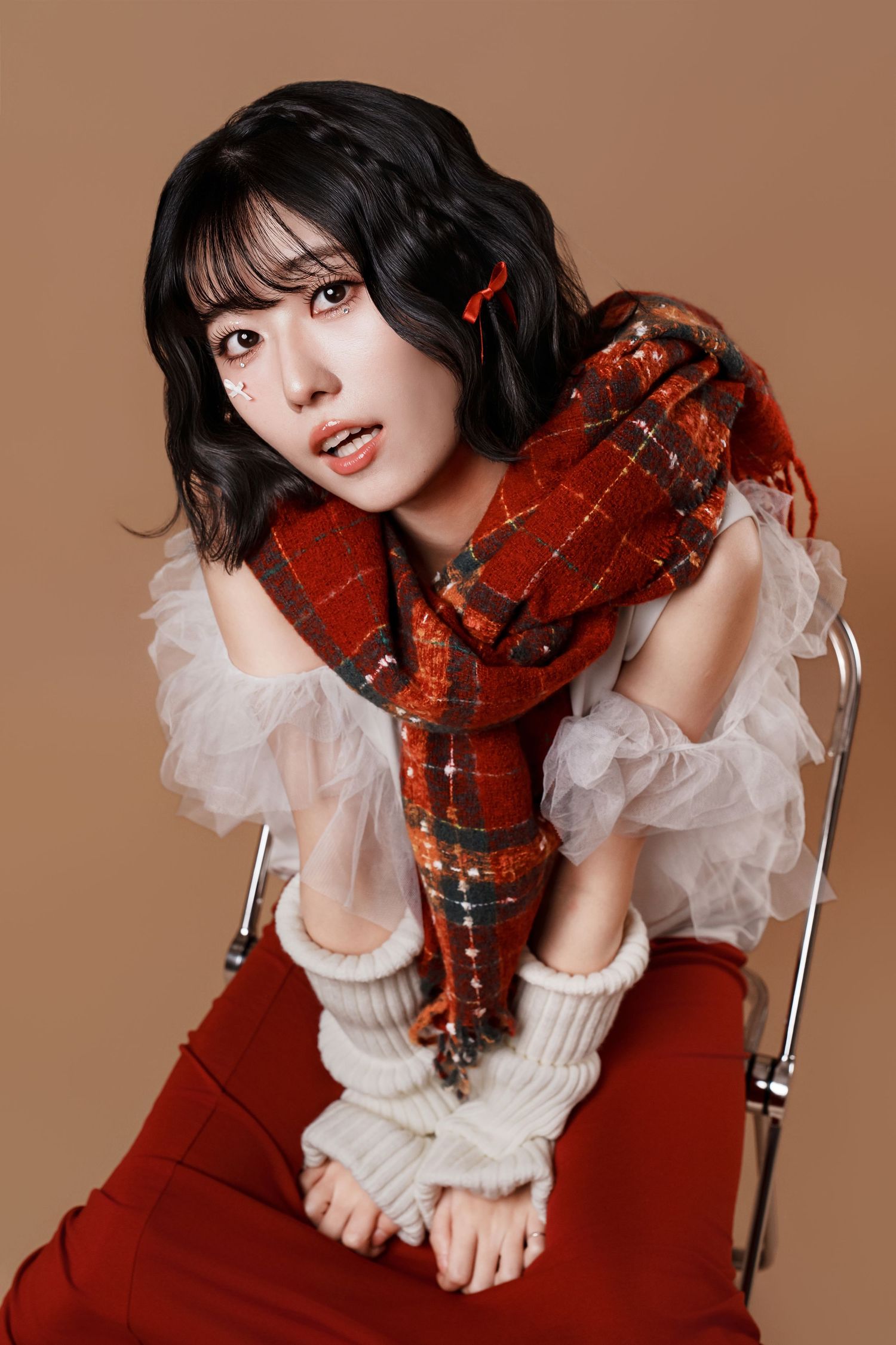A model in a white ruffled blouse and red plaid scarf poses artistically against a brown backdrop.