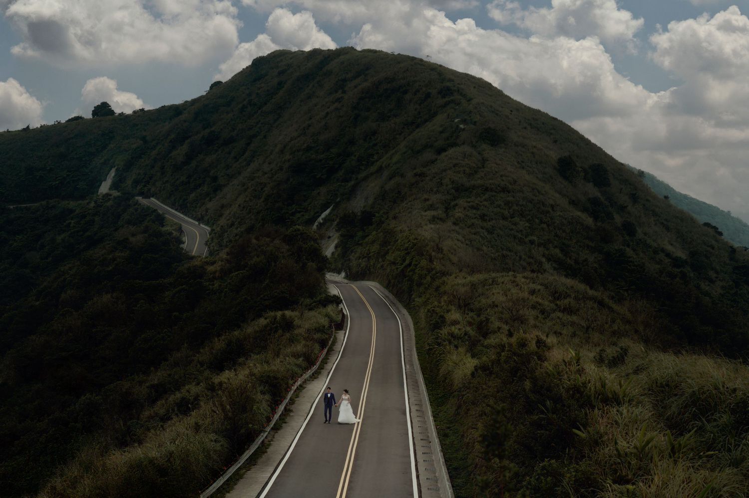Curved mountain highway stretches through rugged terrain under puffy white clouds in a scenic panoramic view.