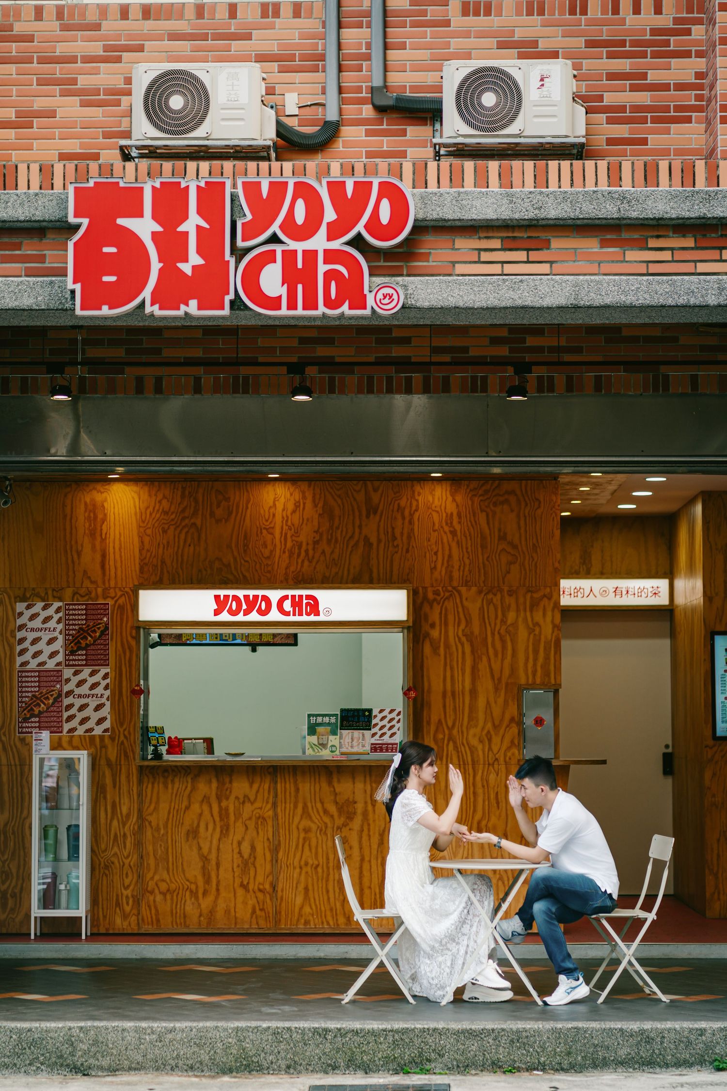 Couple sits at outdoor cafe tables in front of a brick building with Japanese storefront signage.