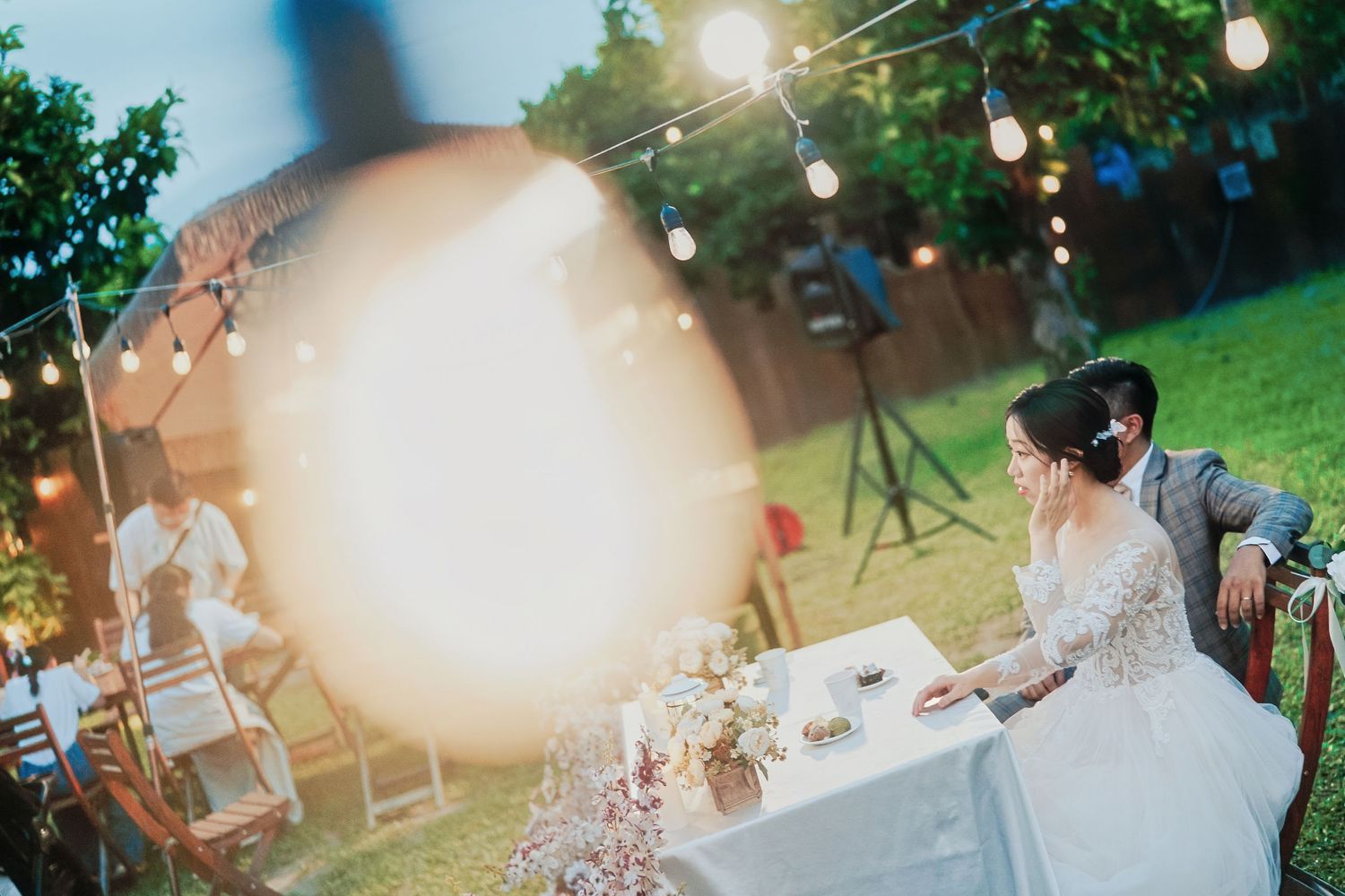 Outdoor wedding reception setup with string lights and table decor during a golden sunset in a garden setting.