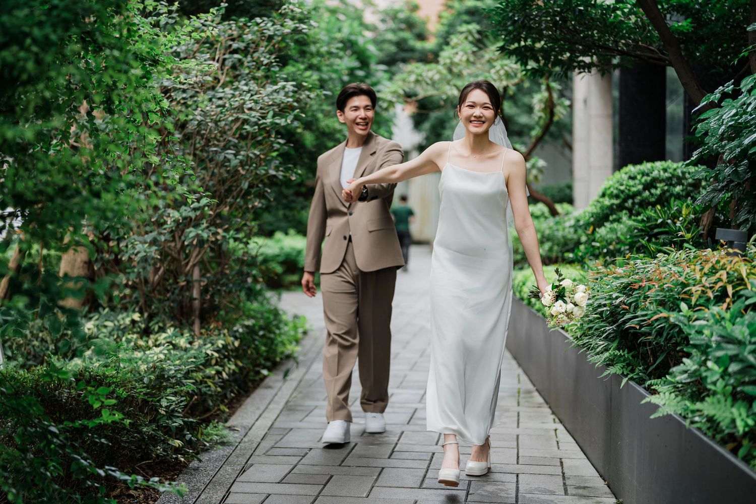 Wedding couple sharing joyful moment while walking along landscaped garden path with stone walls.