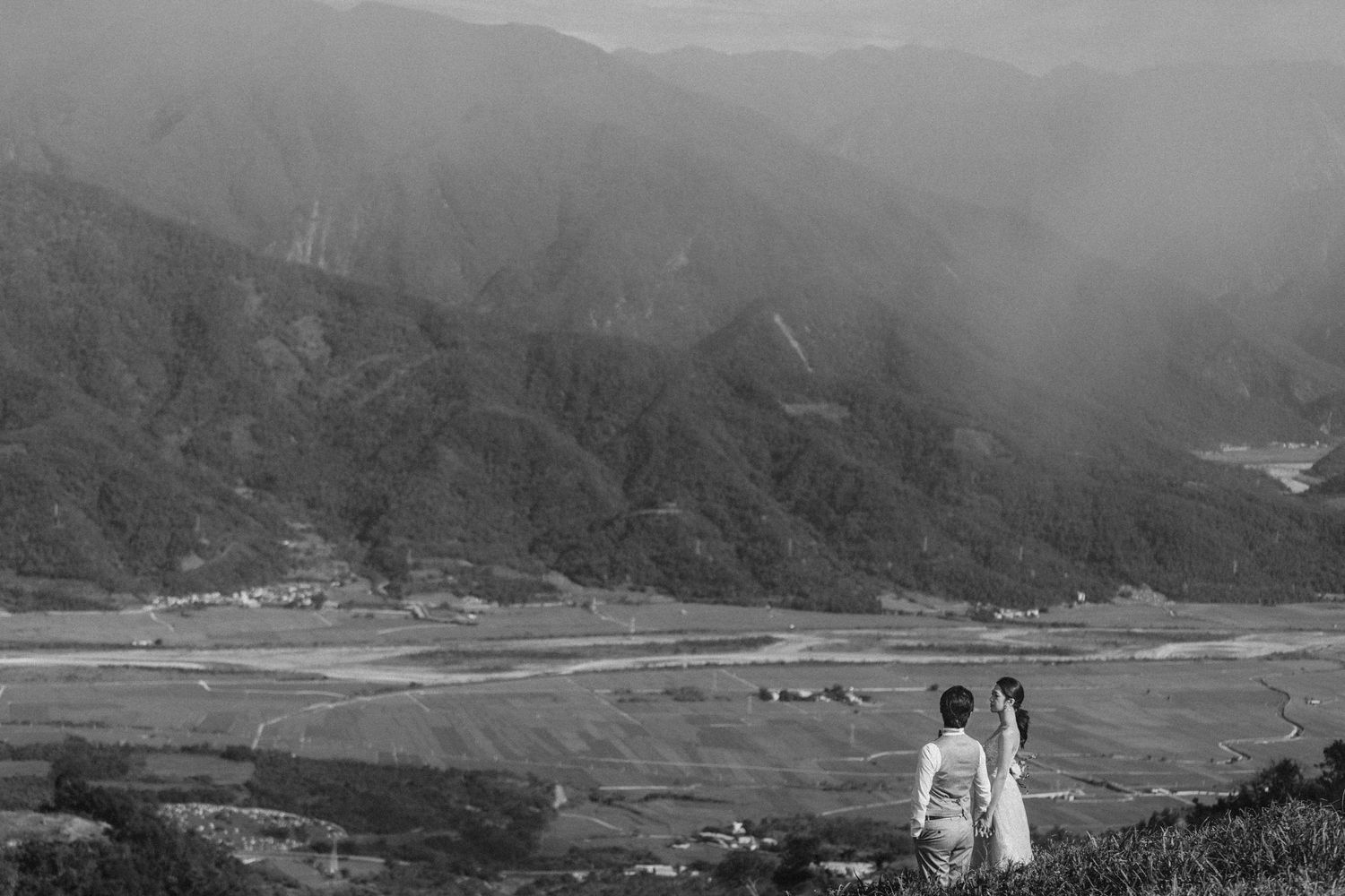 Black and white photo of a couple standing on a hillside overlooking a vast mountain valley landscape.