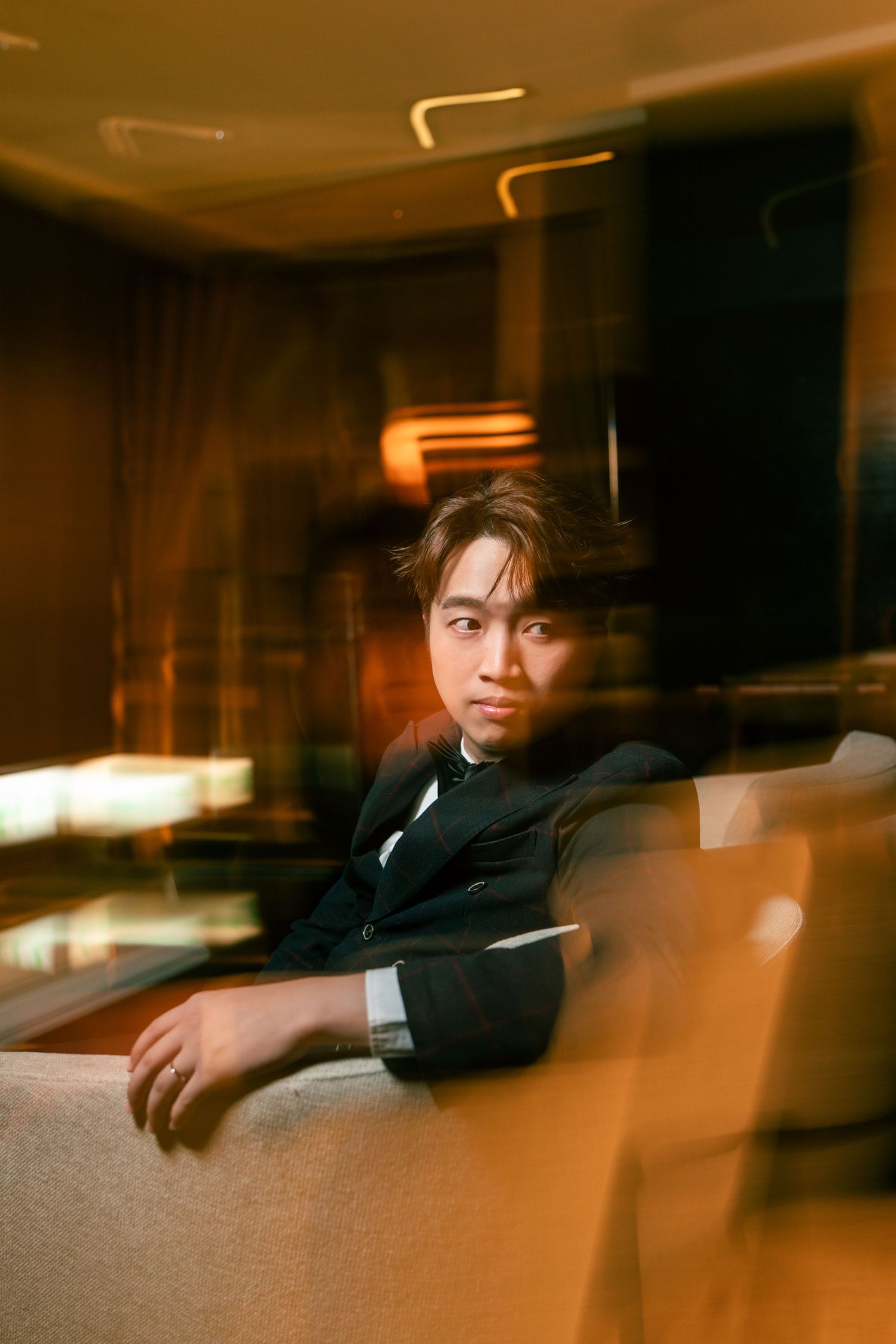 Young man in black suit sitting on sofa in dimly lit room with warm amber lighting and motion blur effect.
