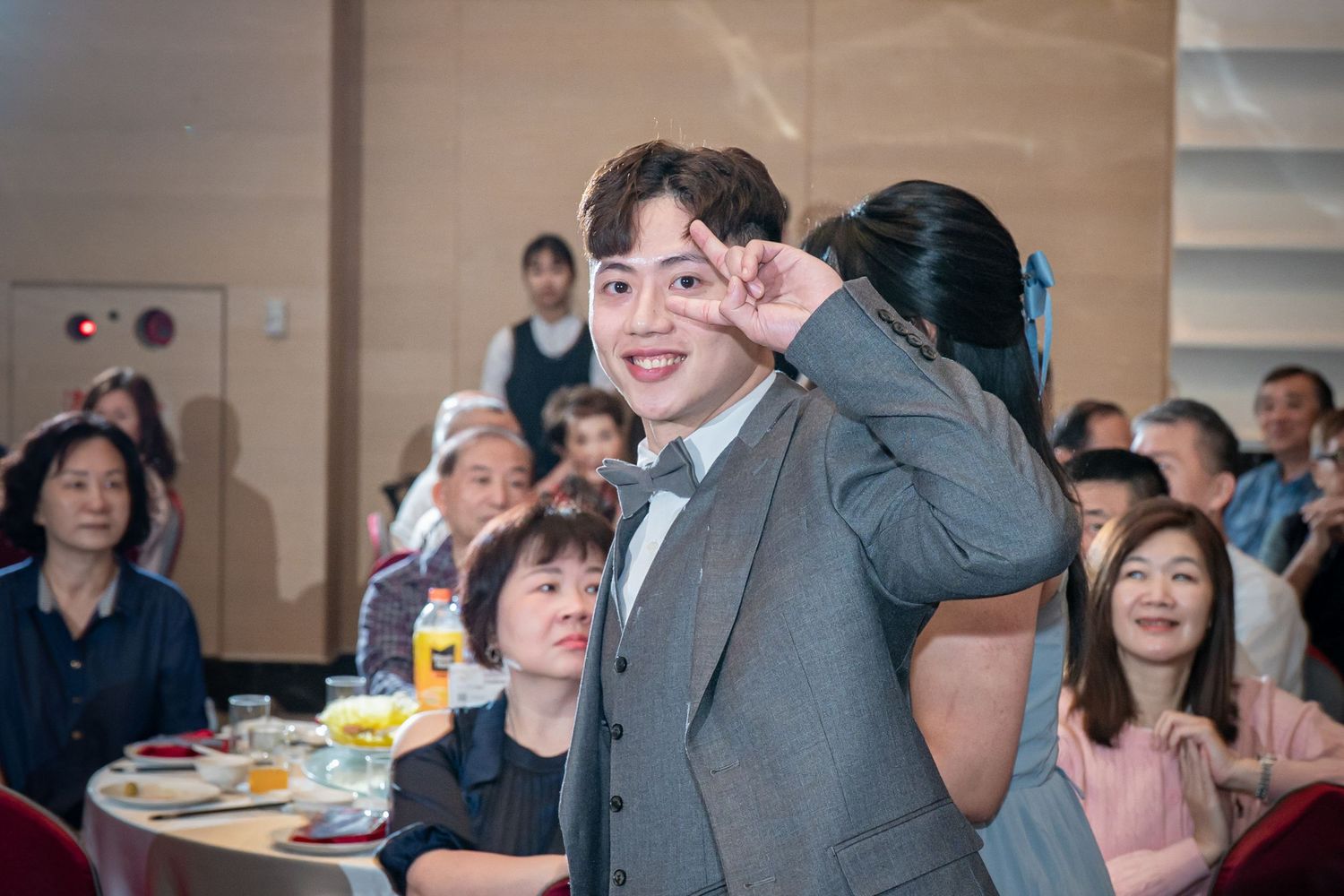Young man in gray suit making peace sign gesture at a formal banquet with guests seated at round tables.