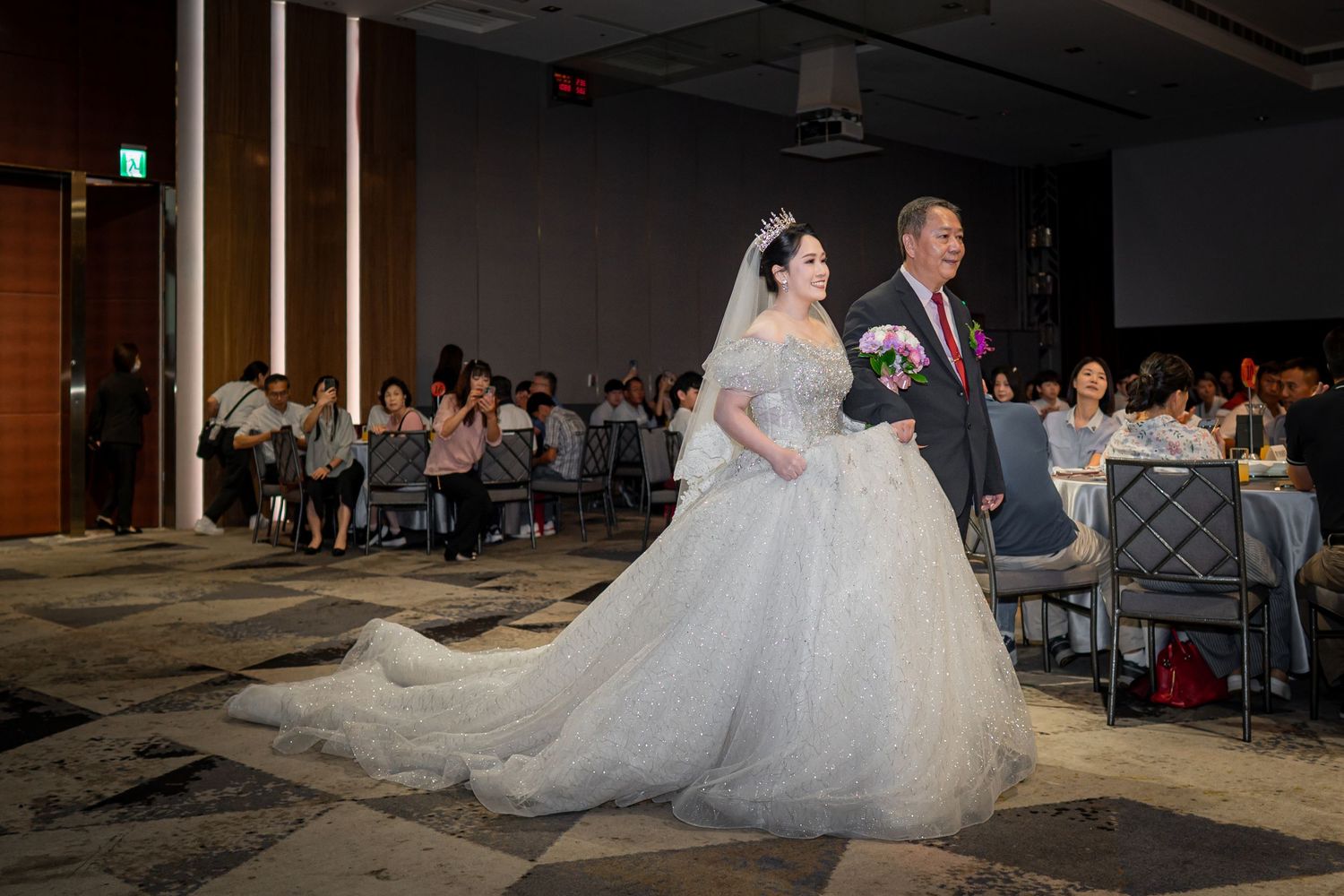 A couple walks down the aisle at a formal wedding reception in an elegant hotel ballroom with carpeted floors and guests.