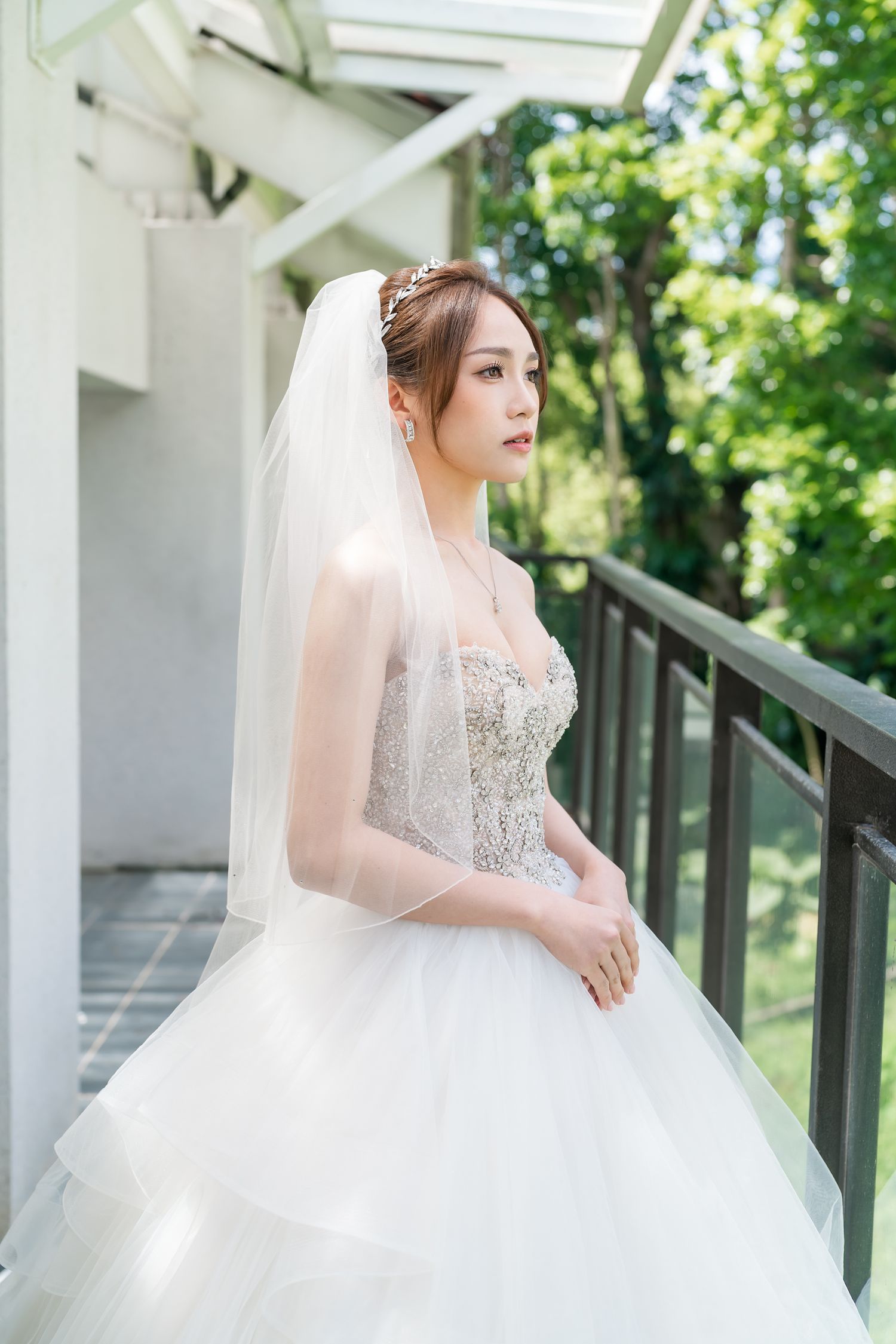 Bride in strapless beaded ballgown and veil poses elegantly on outdoor balcony.