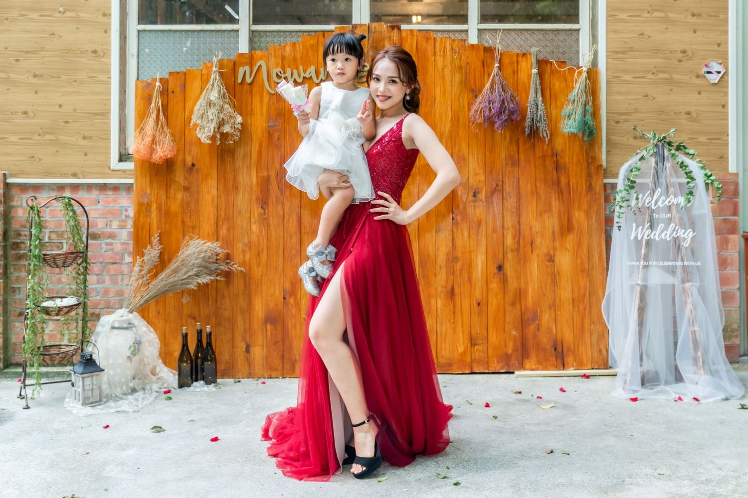 Figure in flowing red gown poses with small child against wooden wall with dried flower decorations.