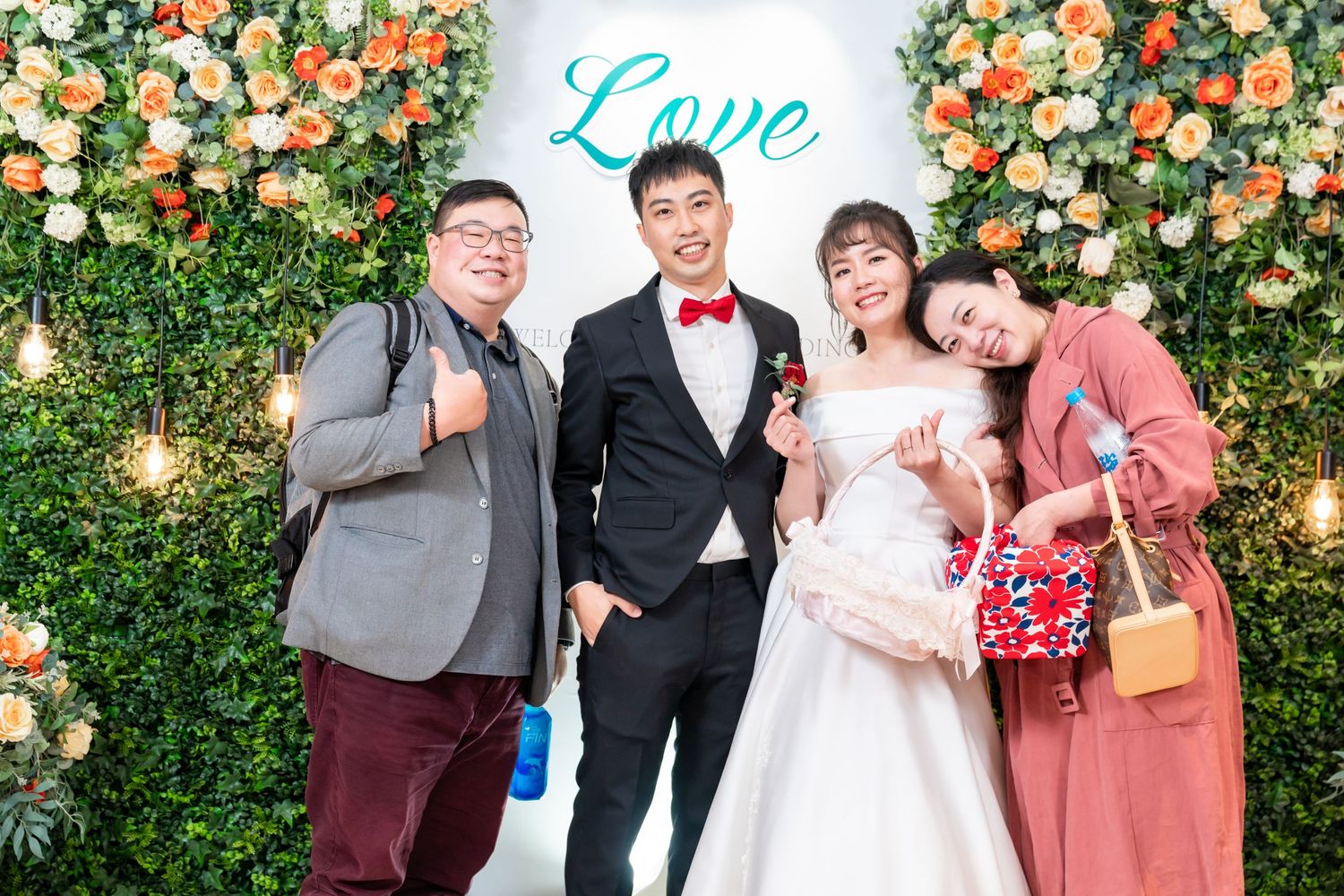 Small wedding group strikes playful poses in front of a floral wall with 'Love' signage.