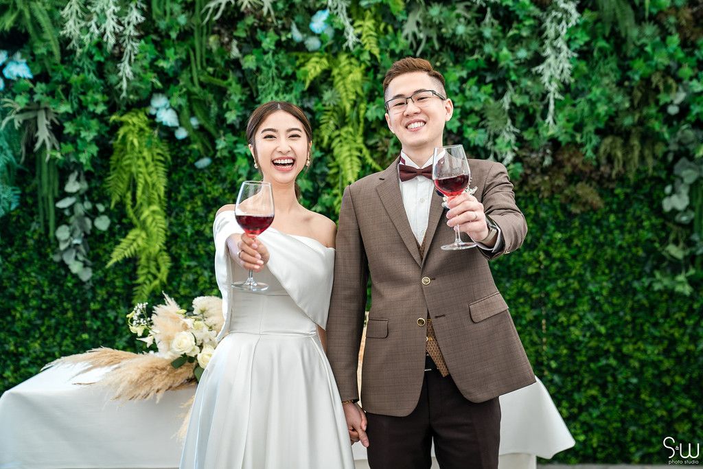 A couple toasts with wine glasses against a green floral backdrop.