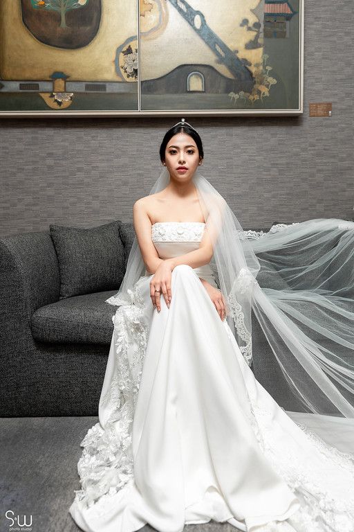 A bride in an elegant white wedding gown poses on a grey sofa with flowing veil.