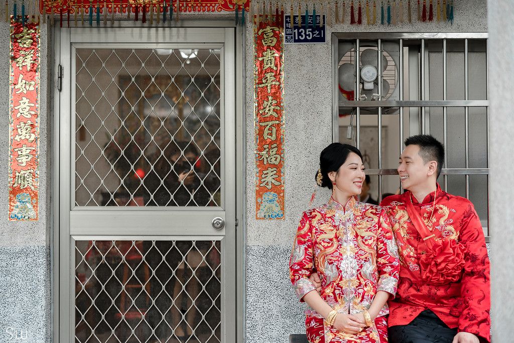 Couple in traditional Chinese wedding attire standing in front of a doorway with red decorations.