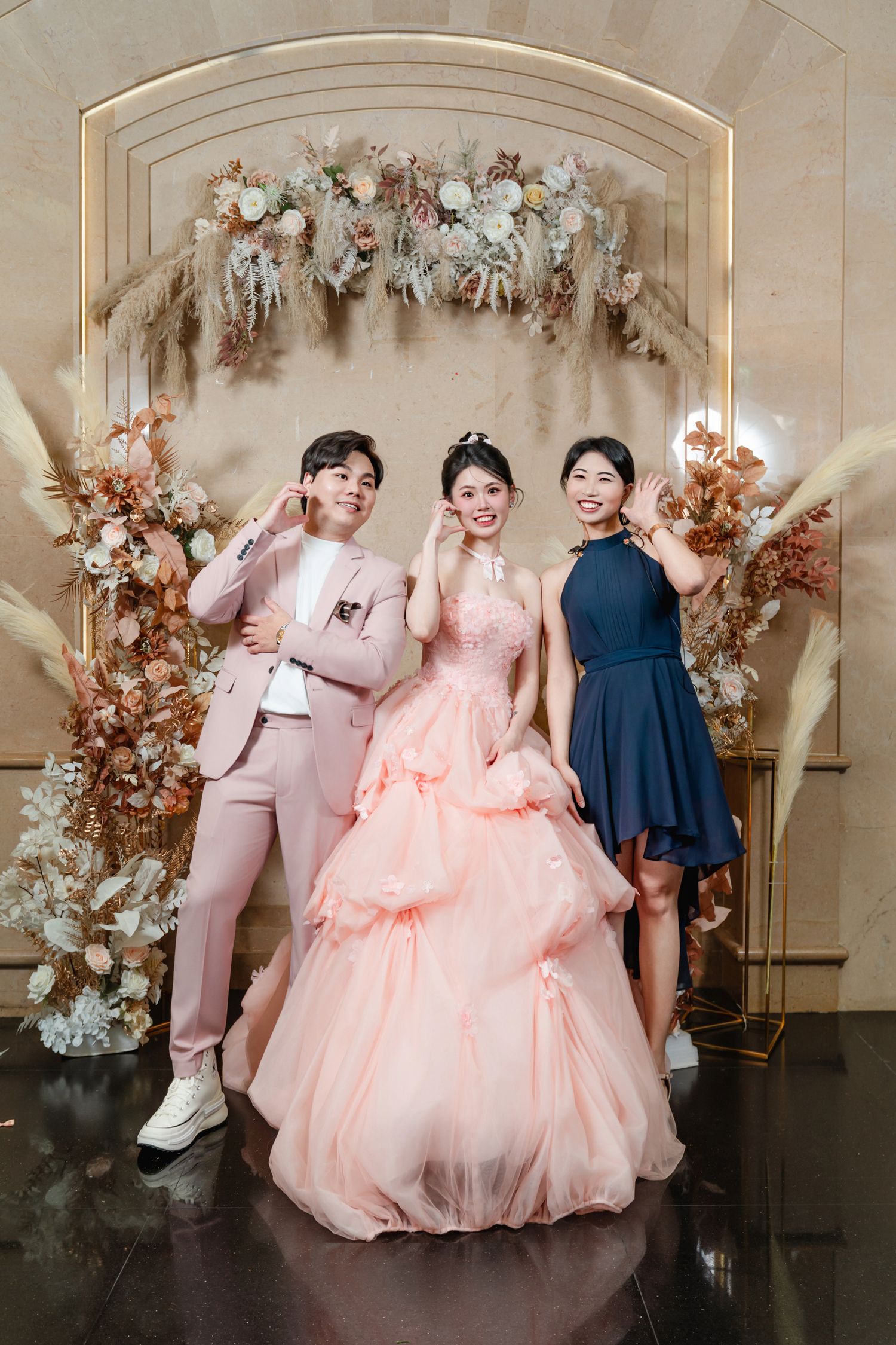 Three people pose in formal attire against an elegant floral arch backdrop with pink and cream flowers and dried pampas grass.