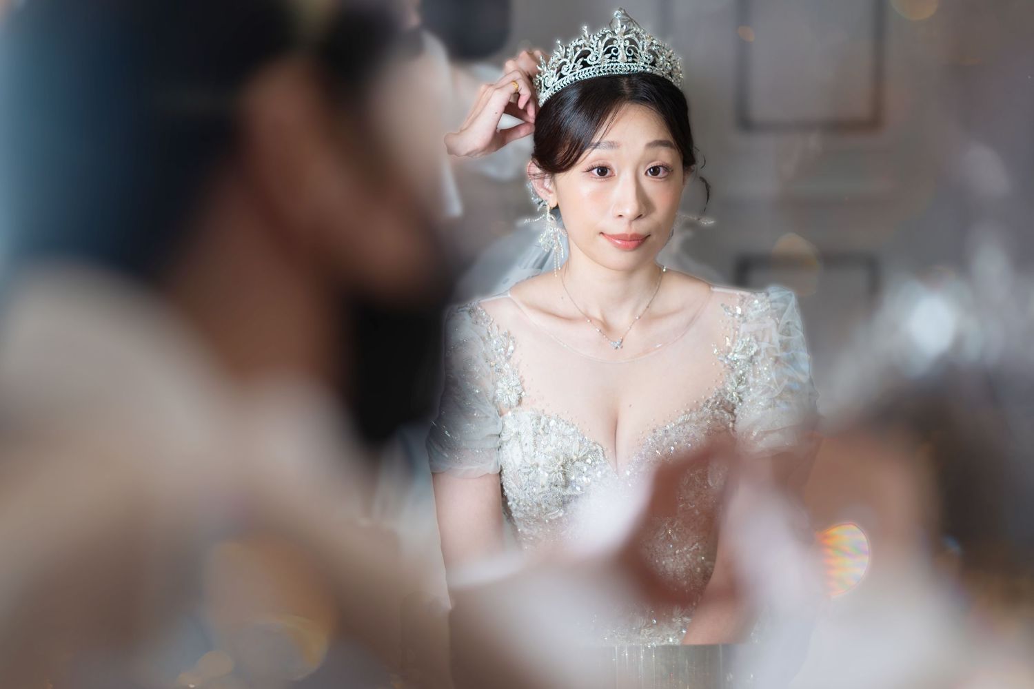 A bride wears a delicate white lace dress with sheer details and a sparkling tiara in an elegant indoor setting.