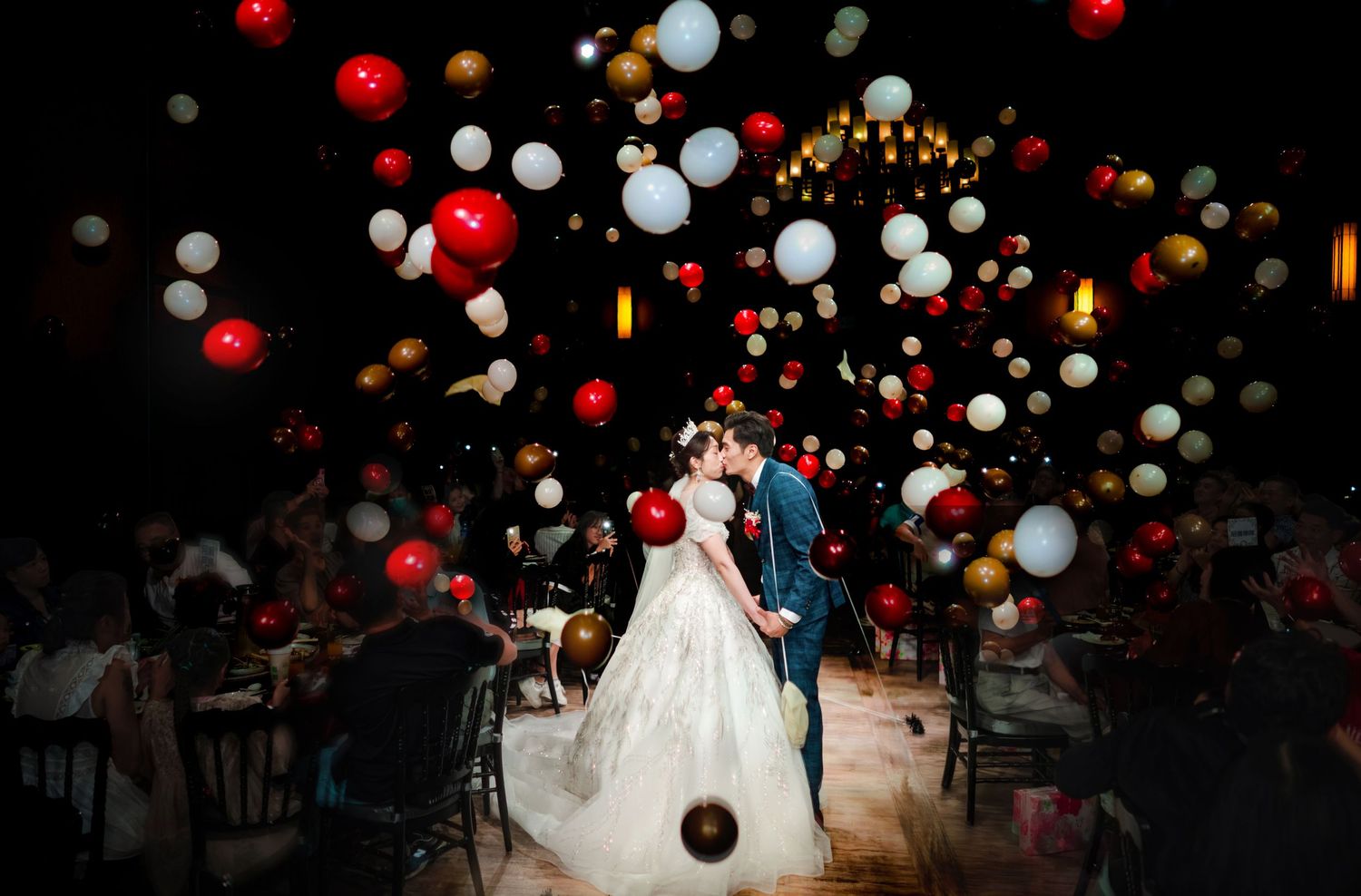 A couple shares their first dance at a wedding reception under floating red and white balloons in a dark venue.