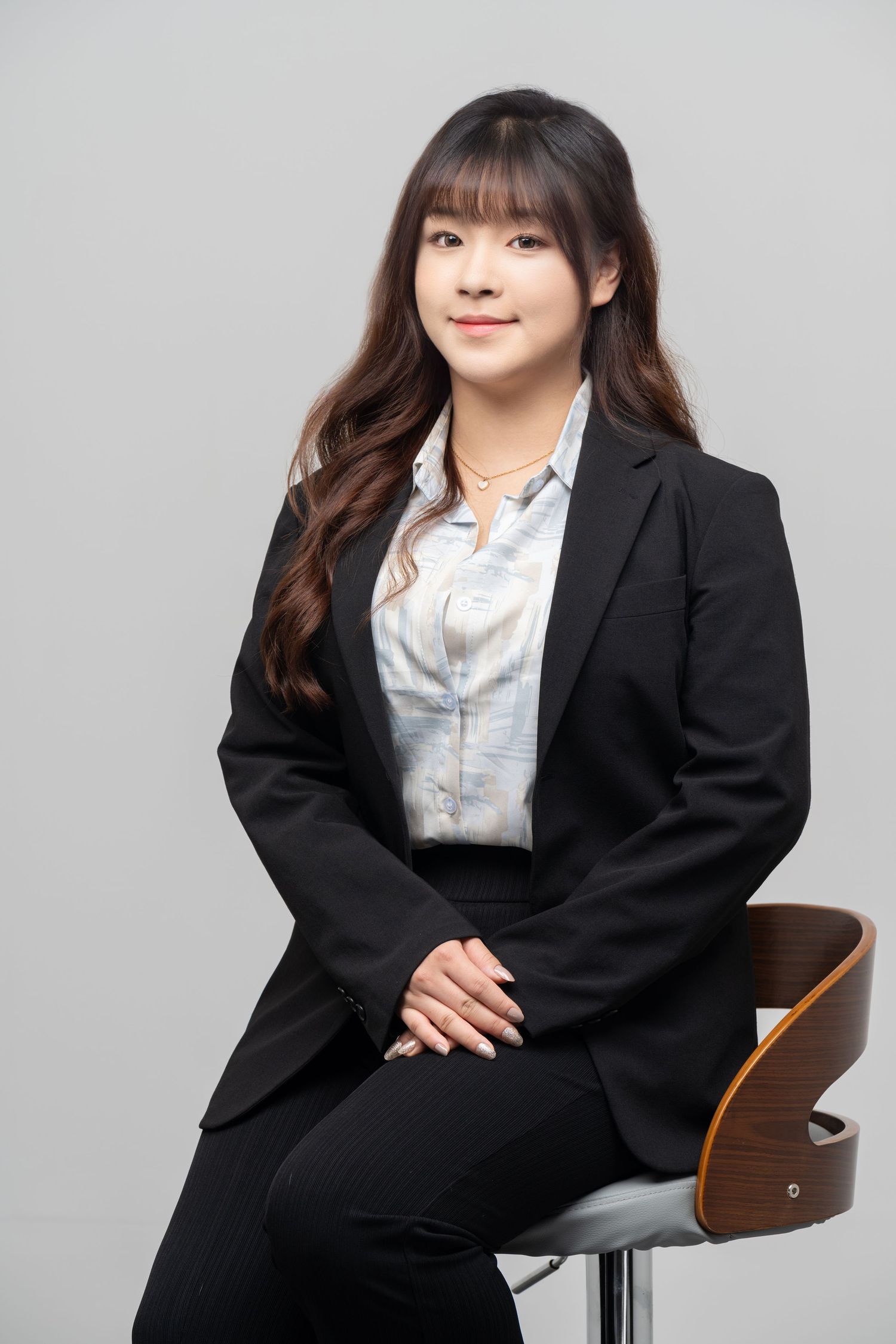 Professional portrait of someone in a black business suit and light colored blouse seated on a modern chair against gray background.