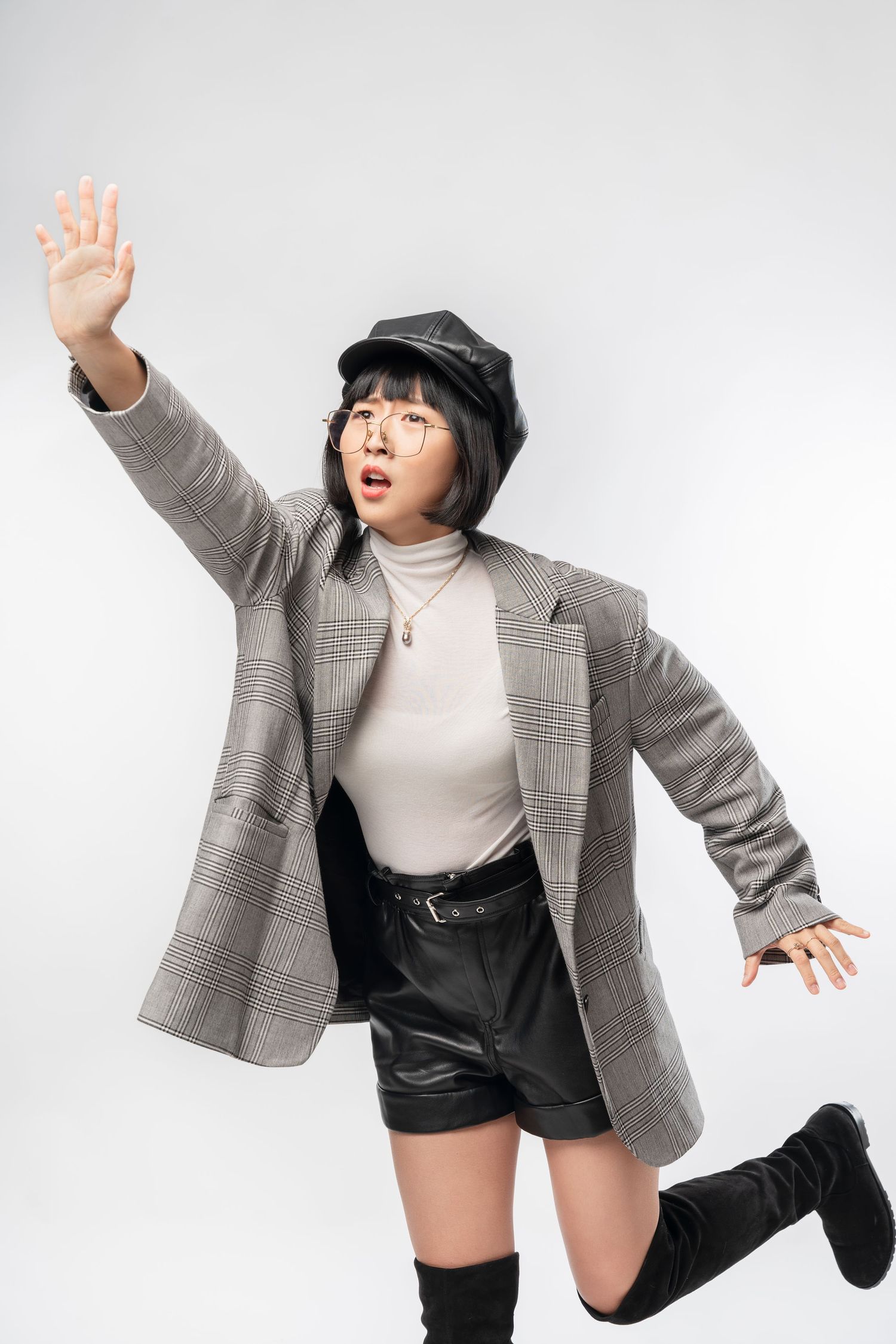 Stylish young woman in plaid blazer and leather shorts strikes dynamic pose wearing black boots and newsboy cap.