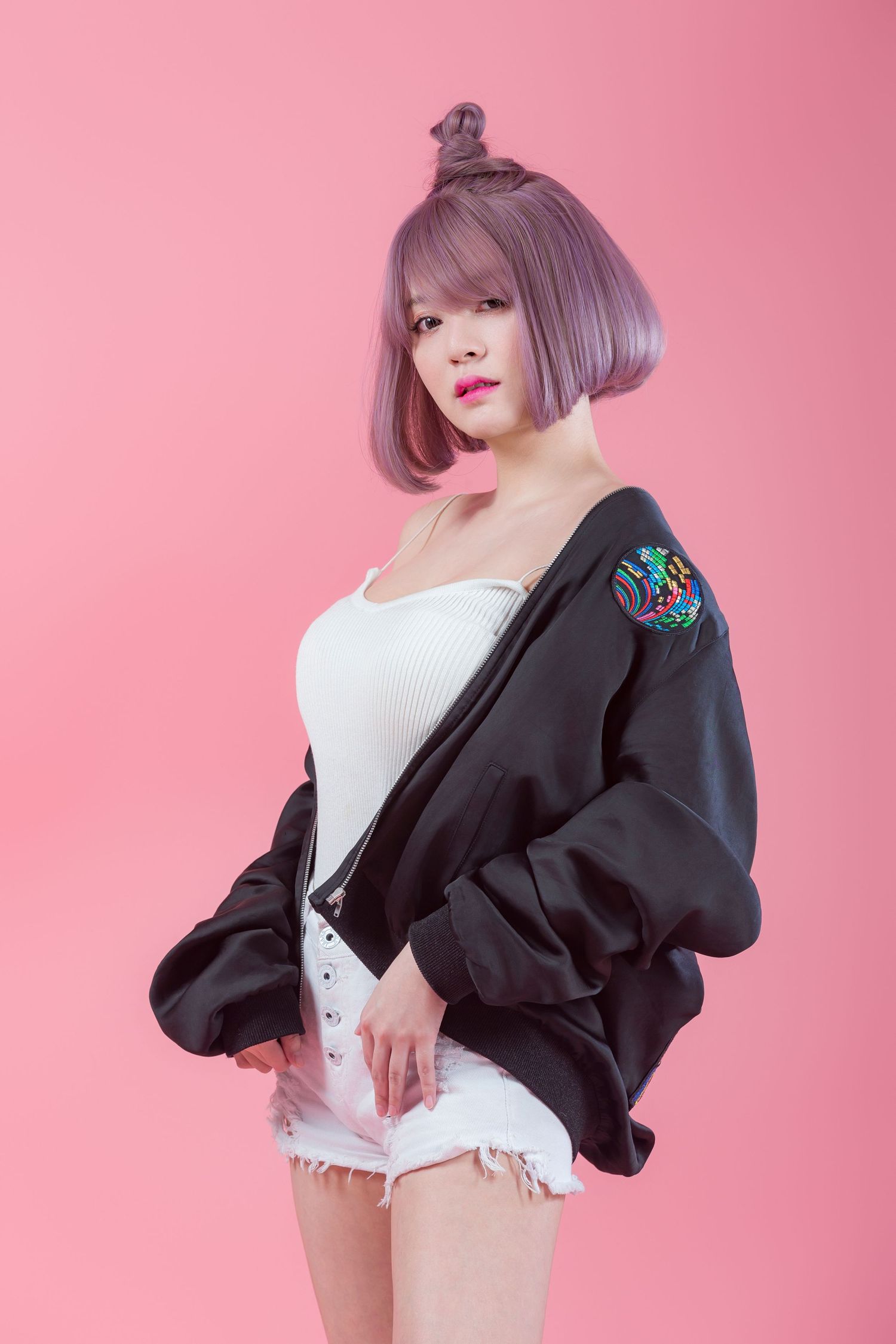 A model in a black varsity jacket and white shorts poses against a pink backdrop, sporting a short lavender hairstyle.