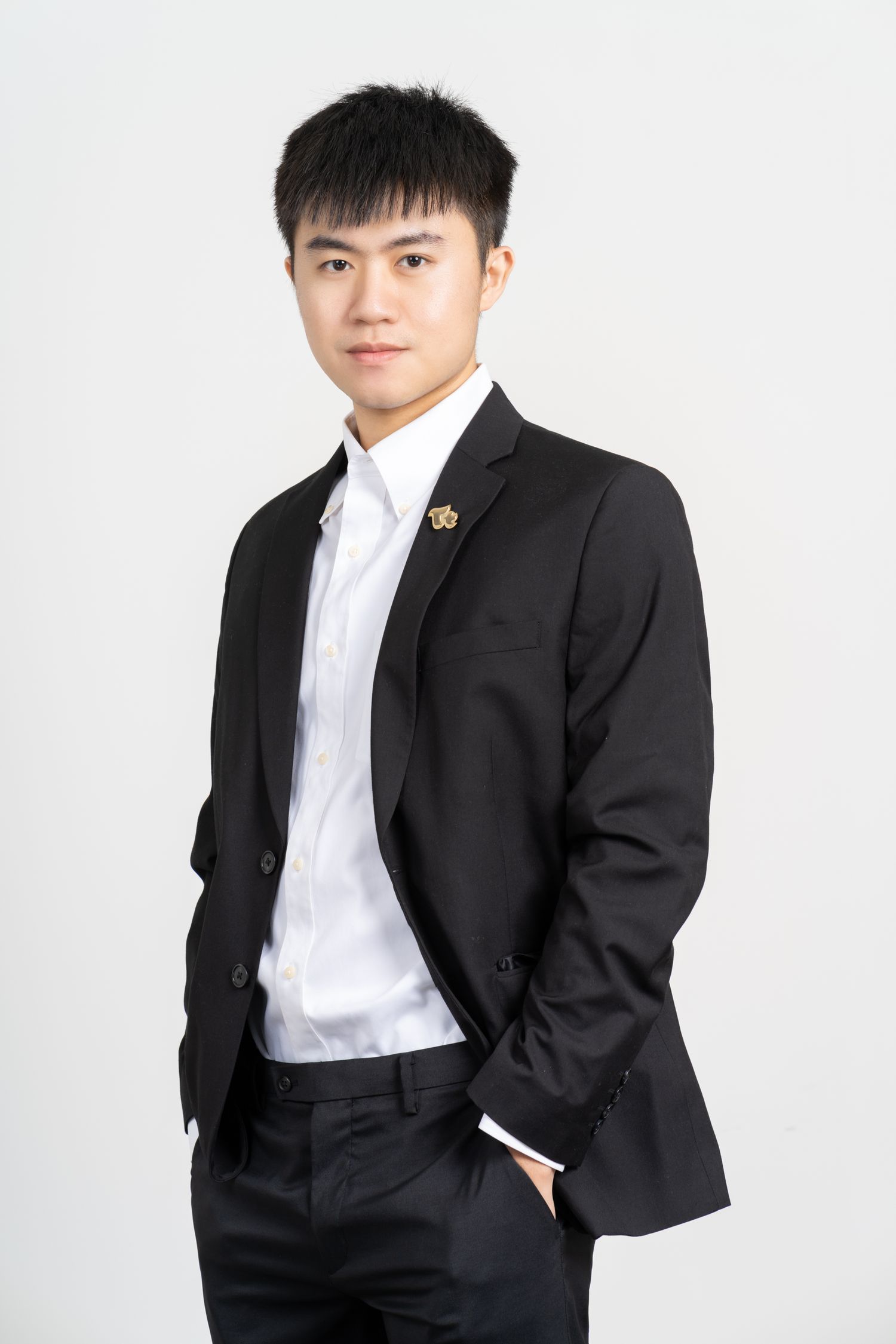 Professional portrait against white background showing someone in a black suit jacket and white dress shirt with lapel pin.