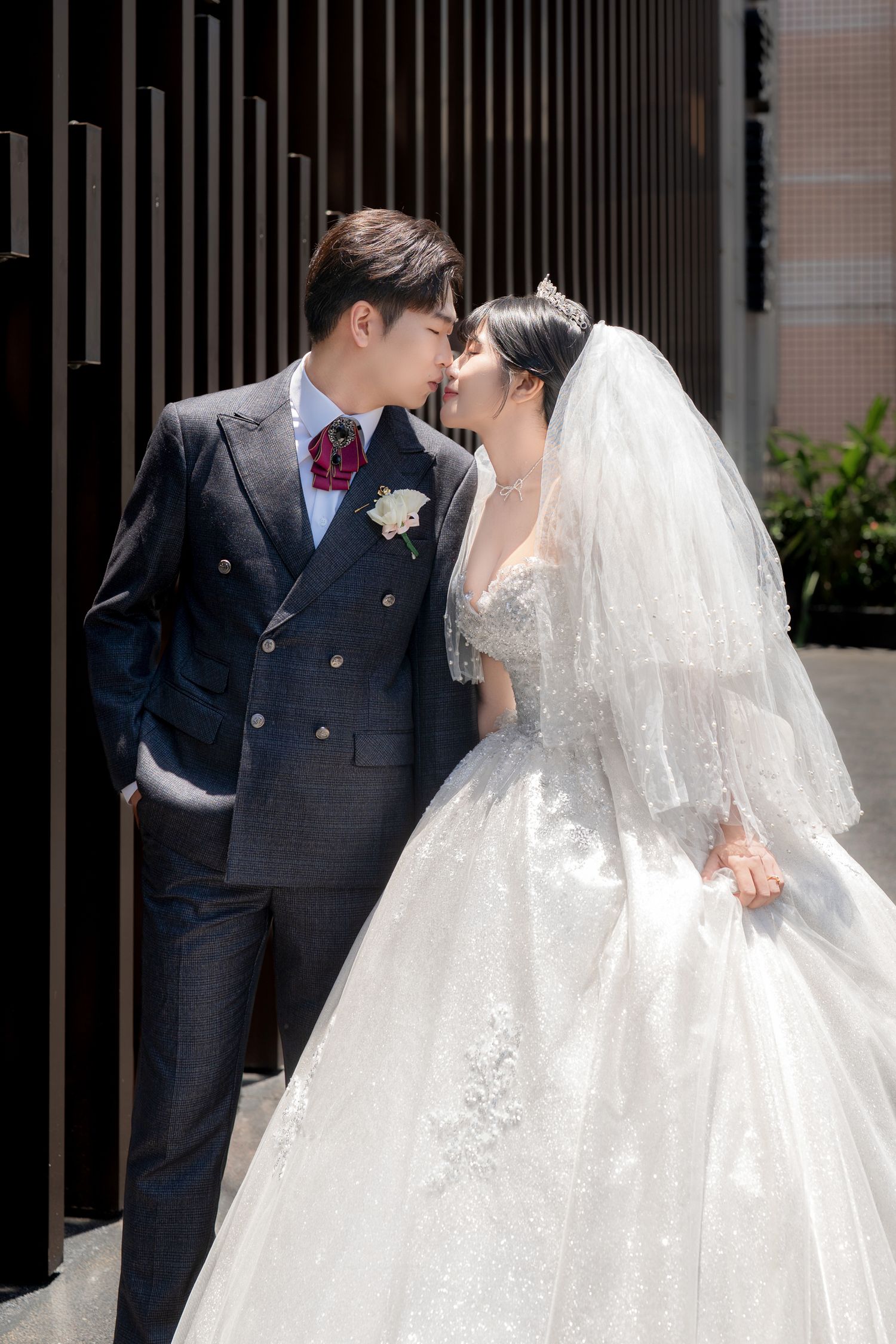 A bride and groom share a romantic moment, nearly kissing outdoors, she in a white ball gown and he in a dark suit.