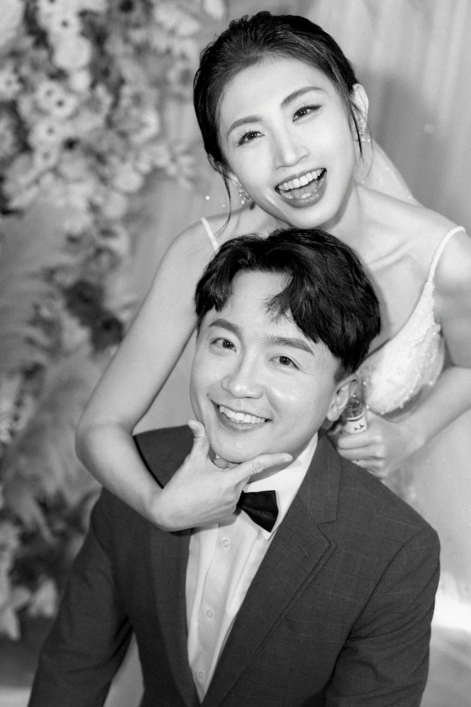 A joyful black and white wedding portrait showing a couple in formal attire smiling against a floral backdrop.