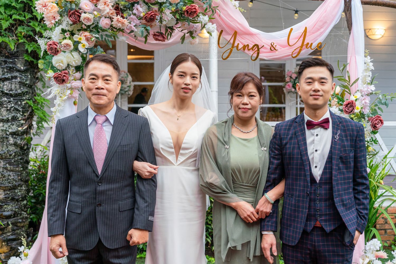 A group of four people posing under a floral arch and signage at a wedding celebration.