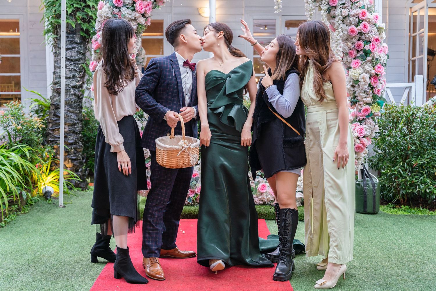 A group gathers outdoors for a celebratory photo on a red carpet with pink floral decorations and greenery in the background.