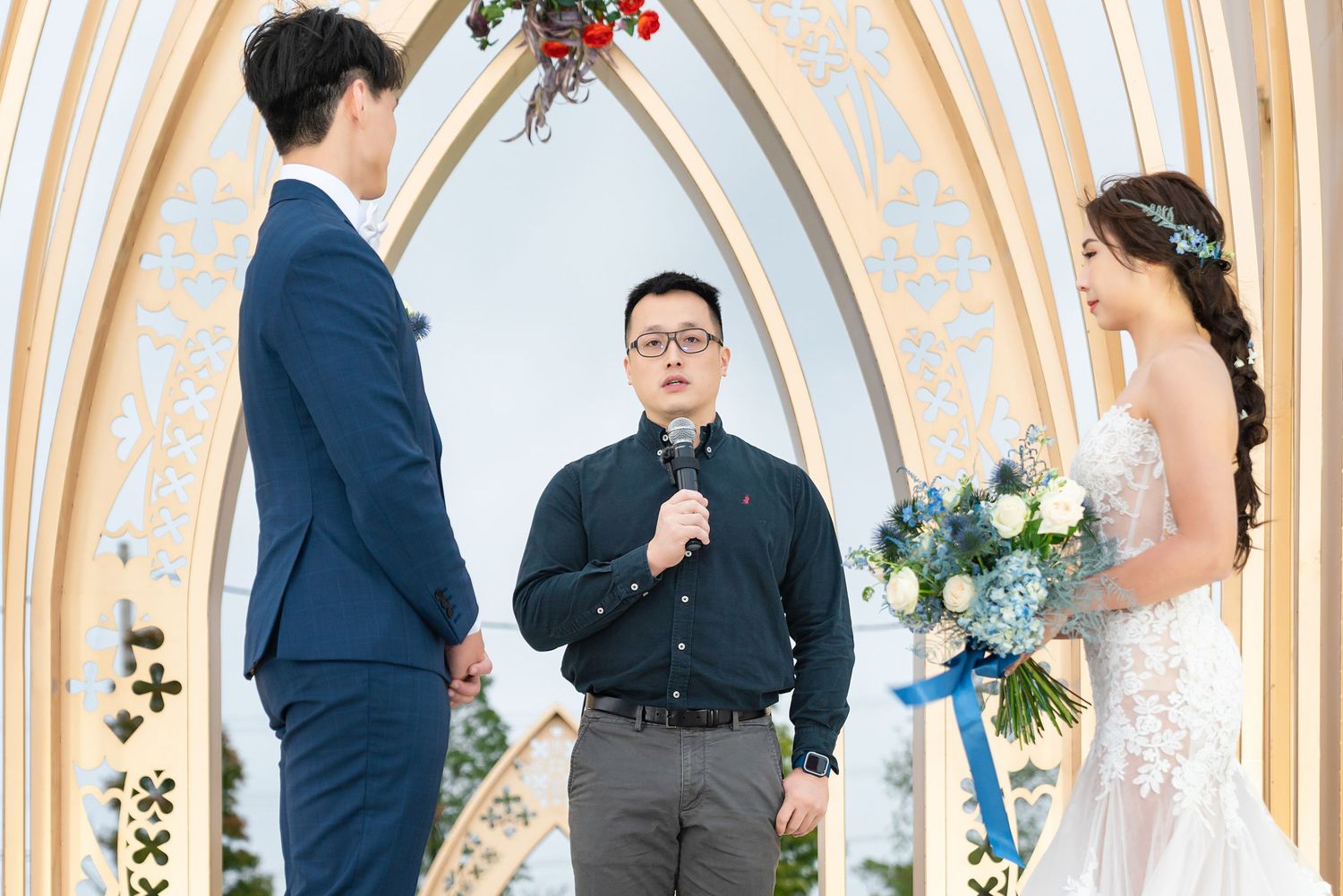 A wedding ceremony takes place under an elegant arched chapel backdrop decorated with flowers and greenery.