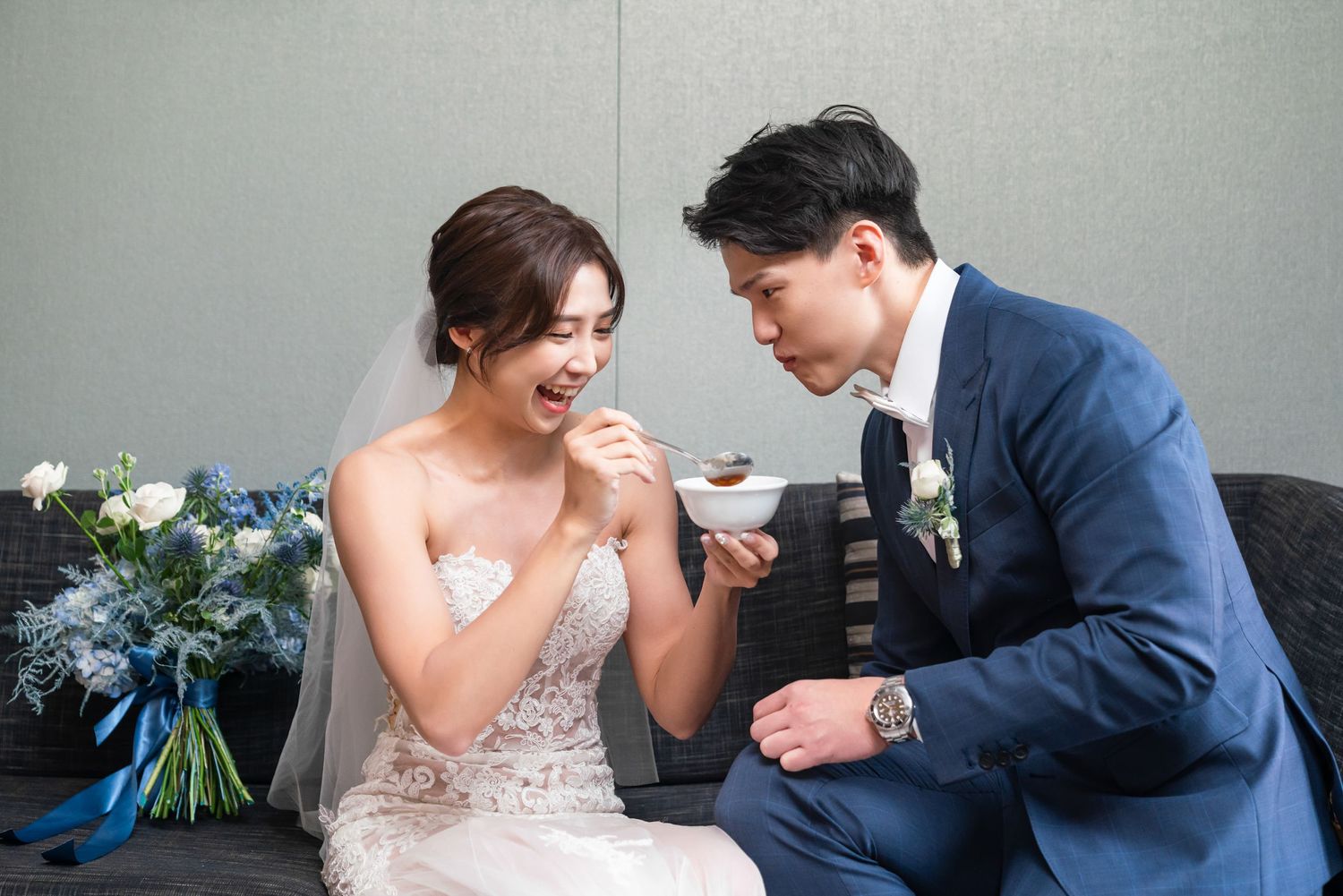 A couple in wedding attire sharing a joyful moment while sitting together on a couch with a floral bouquet nearby.