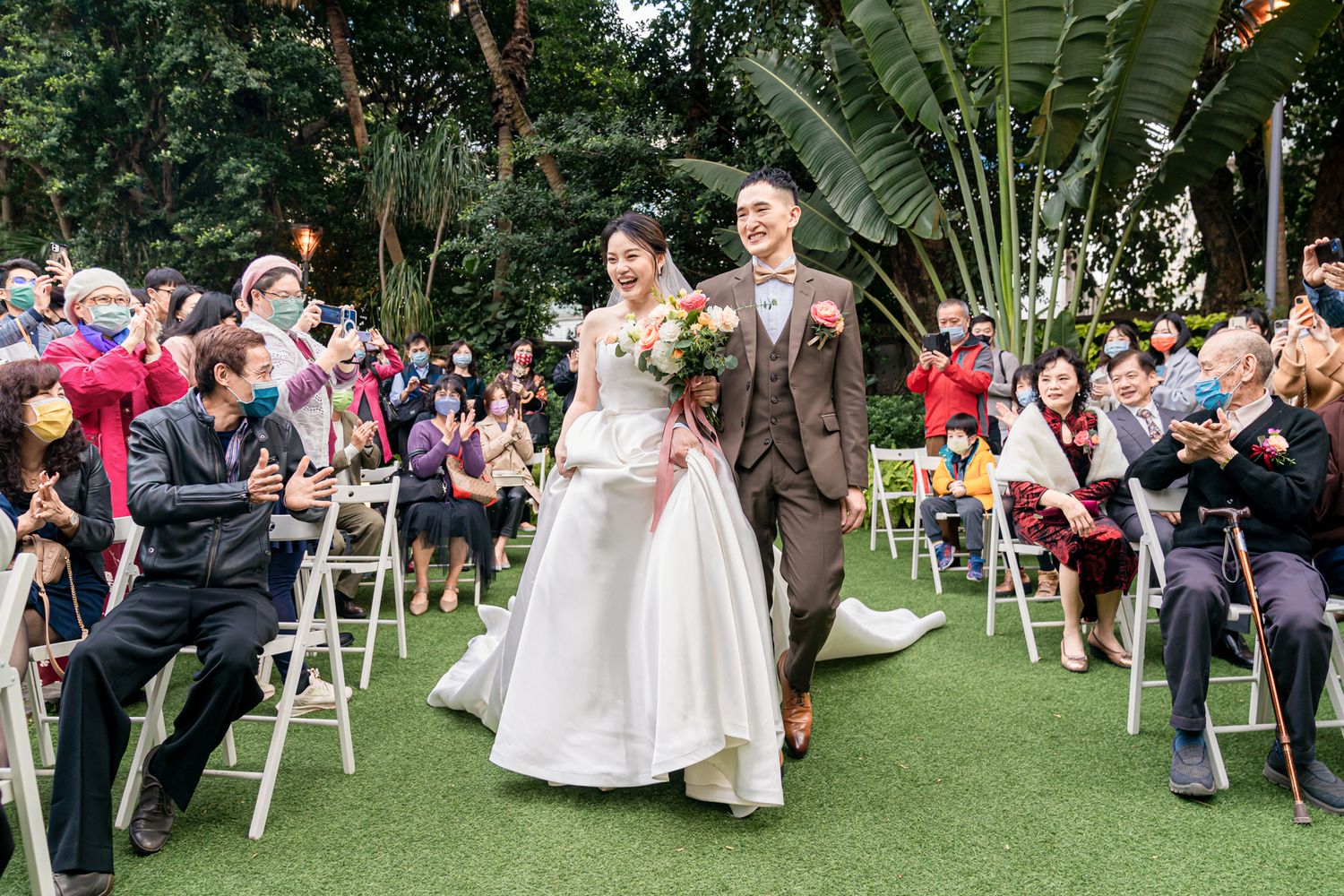 A couple walks down a garden wedding aisle surrounded by guests seated on white chairs and lush tropical foliage.