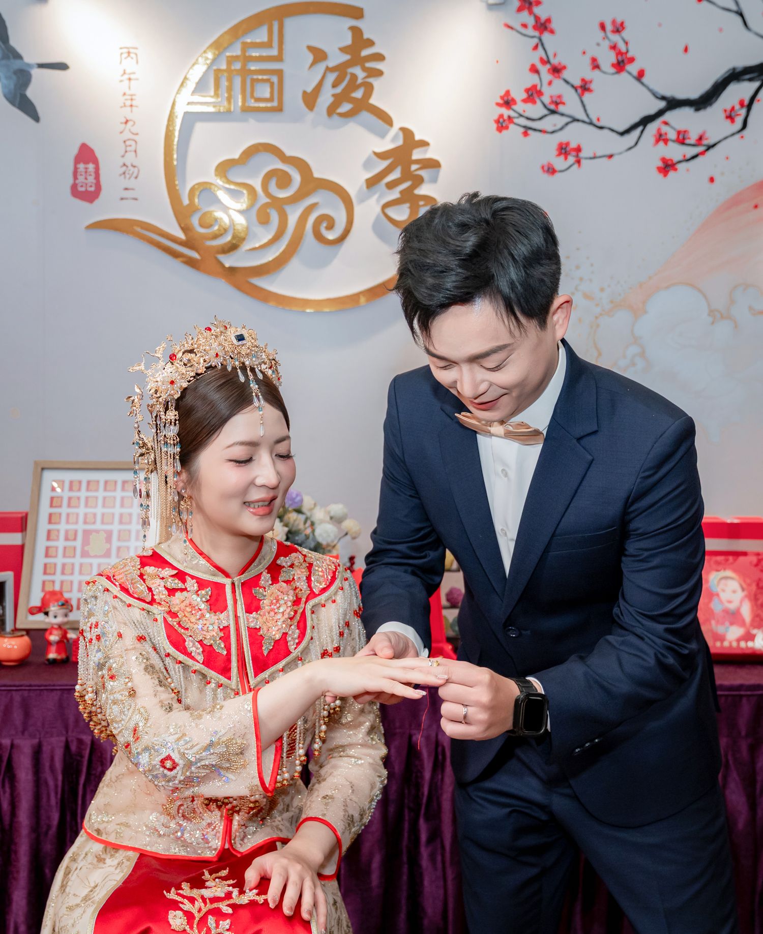 Groom in a navy suit and bow tie places a ring on a bride in an ornate red and gold traditional Chinese wedding gown and headdress.