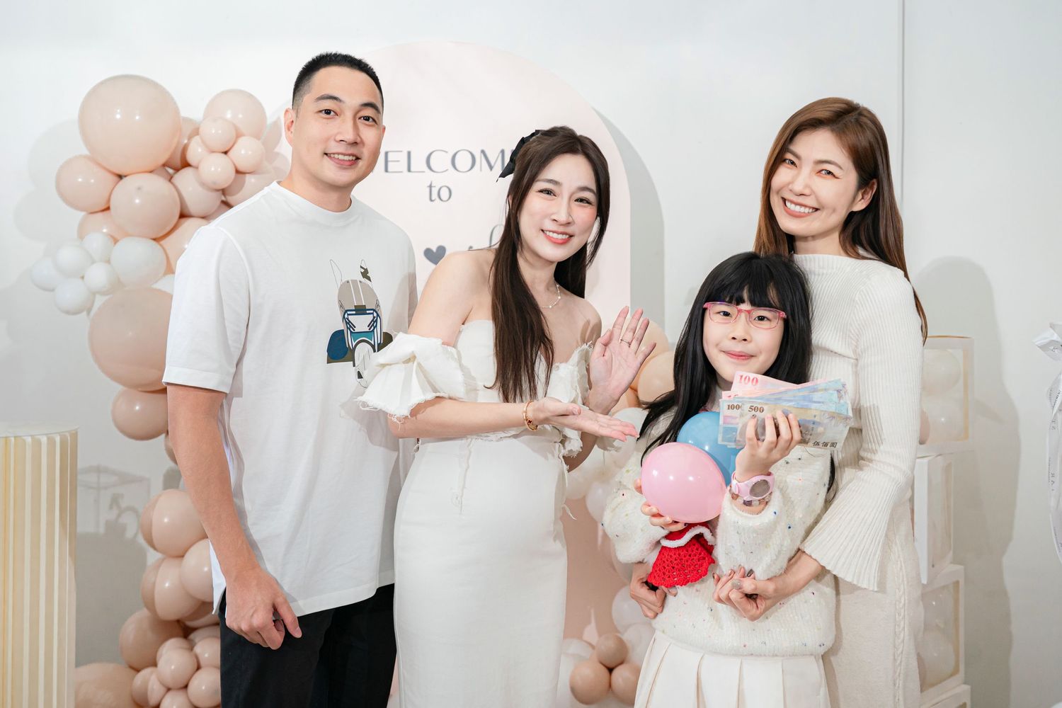 A family in white outfits poses together with pink balloon decorations in a bright studio setting.