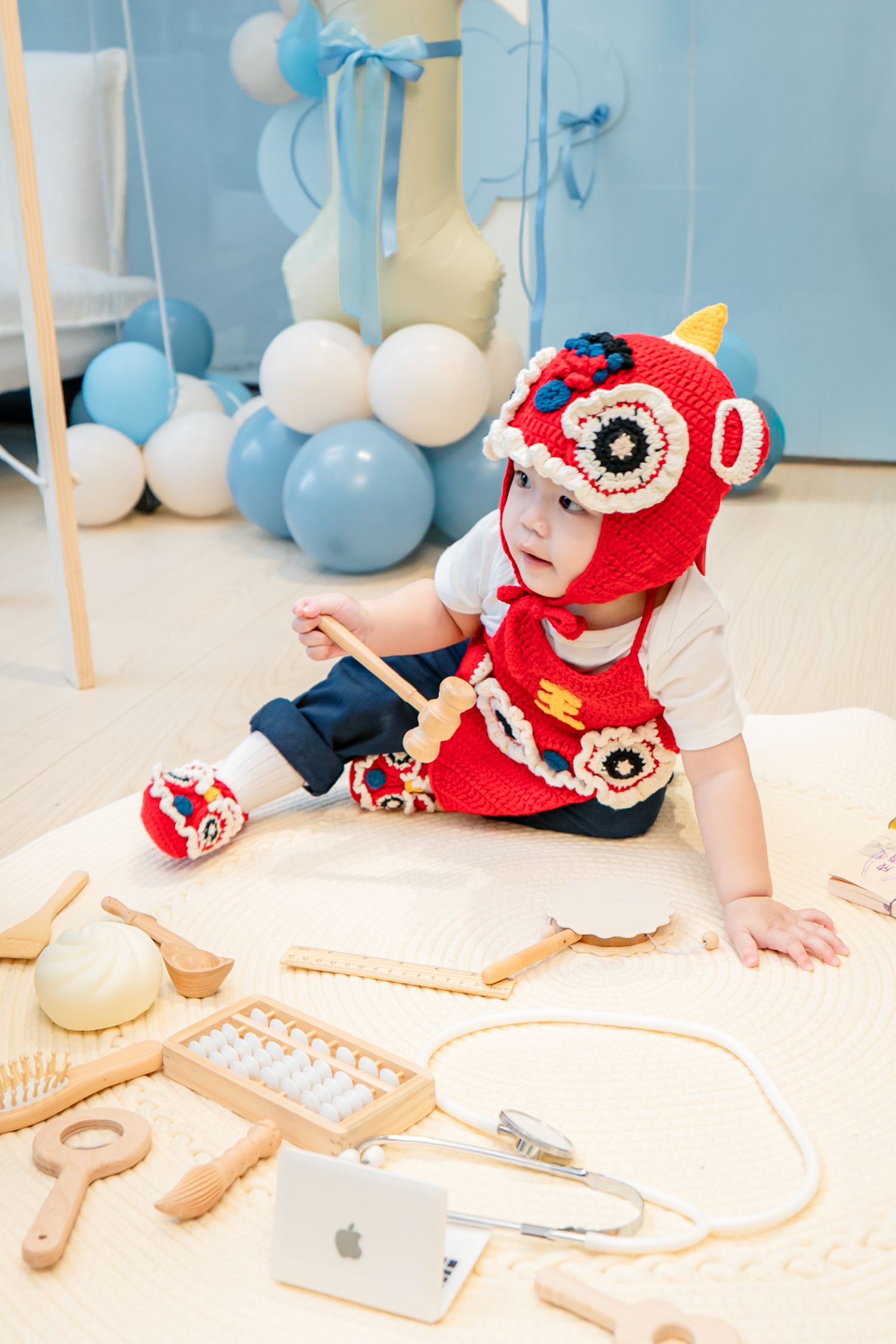 Baby wearing red crochet hat lies on floor surrounded by medical equipment and toys in blue-themed room.