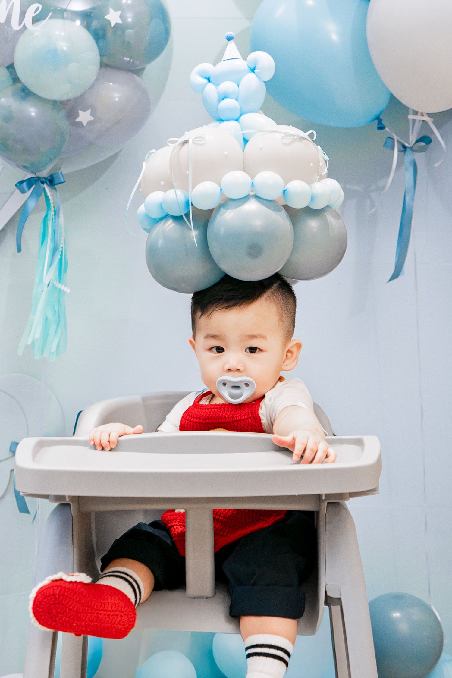 Baby in high chair with balloon crown decoration at blue and white birthday party celebration.